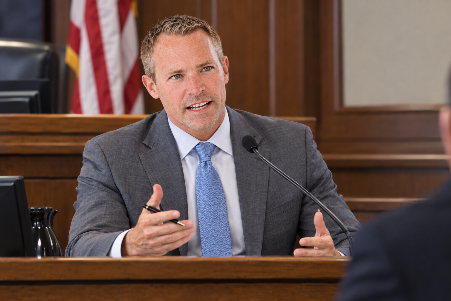 A man in a gray suit and blue tie speaking into a microphone during a formal meeting in a courtroom or government setting, with the American flag in the background.  Dr. Joos testifies on a medical expert ENT case.