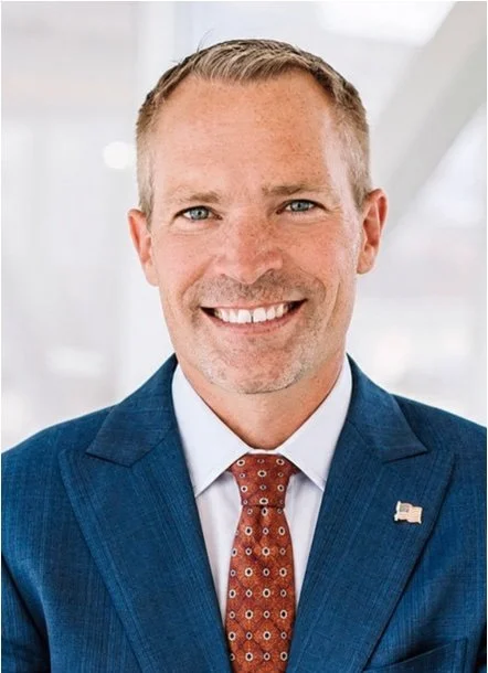 Smiling man wearing a blue suit, white shirt, and red patterned tie, with an American flag lapel pin.  Dr. nathan Joos, a medical expert in the field of otolaryngology.