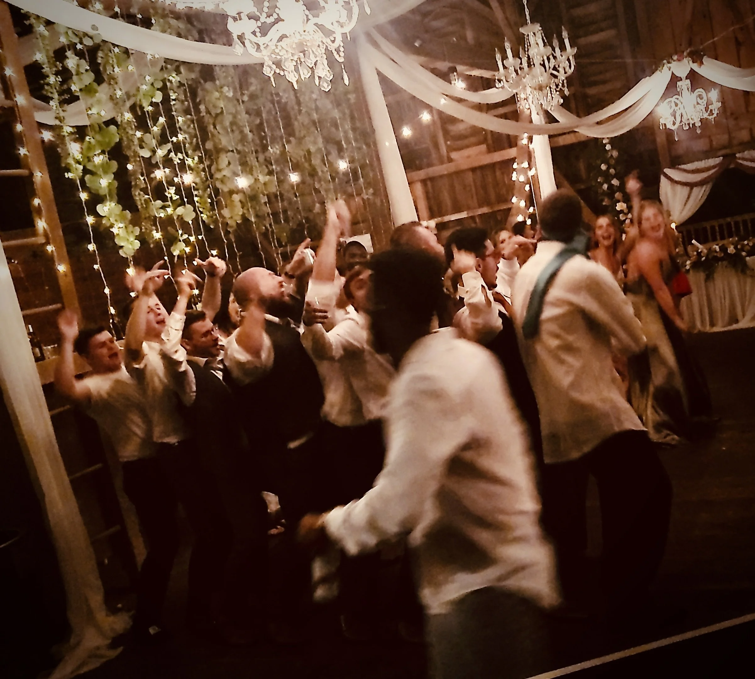 A joyful group of people celebrating at a decorated indoor event with chandeliers, string lights, and draped fabric. Many individuals are smiling, and some are raising their hands in excitement.