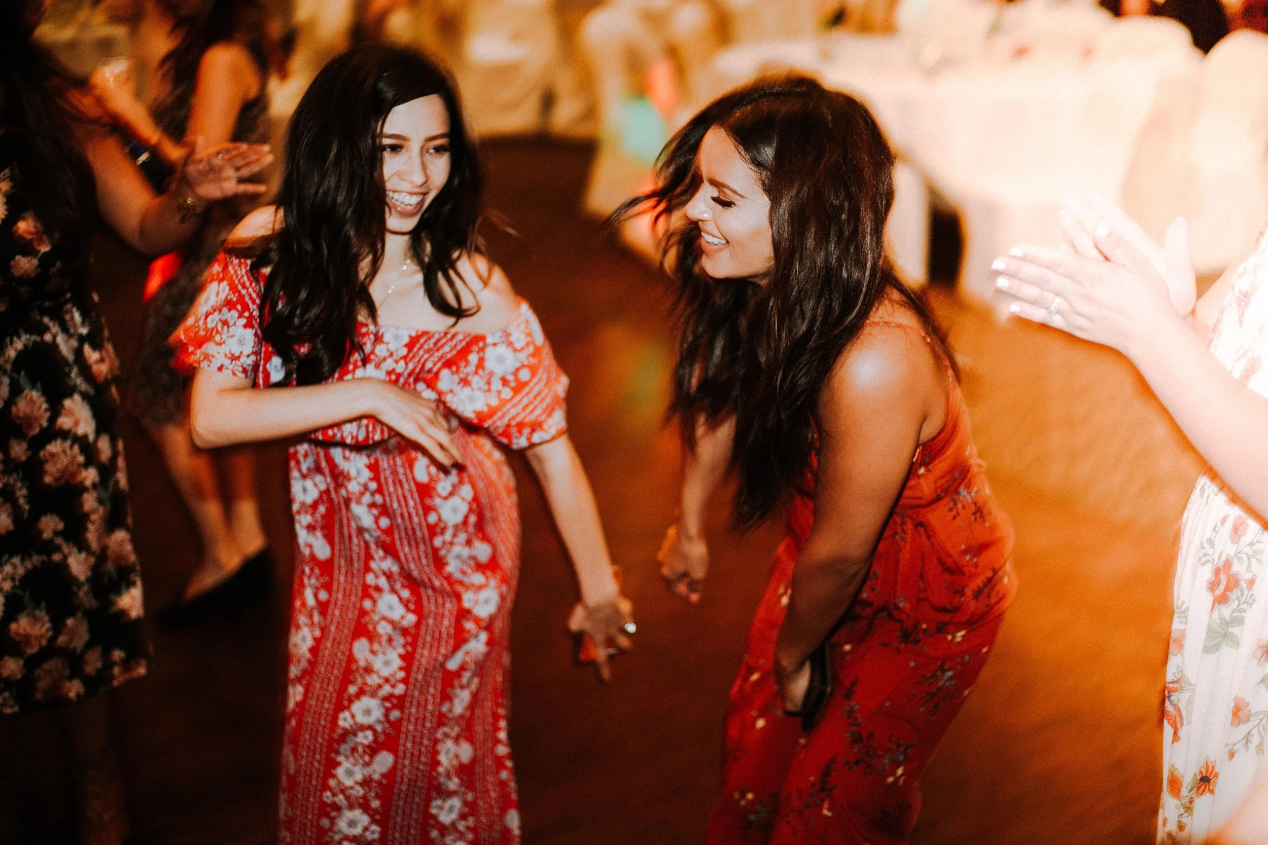 Two women dancing and holding hands at a social event, smiling and enjoying themselves, wearing colorful patterned dresses.