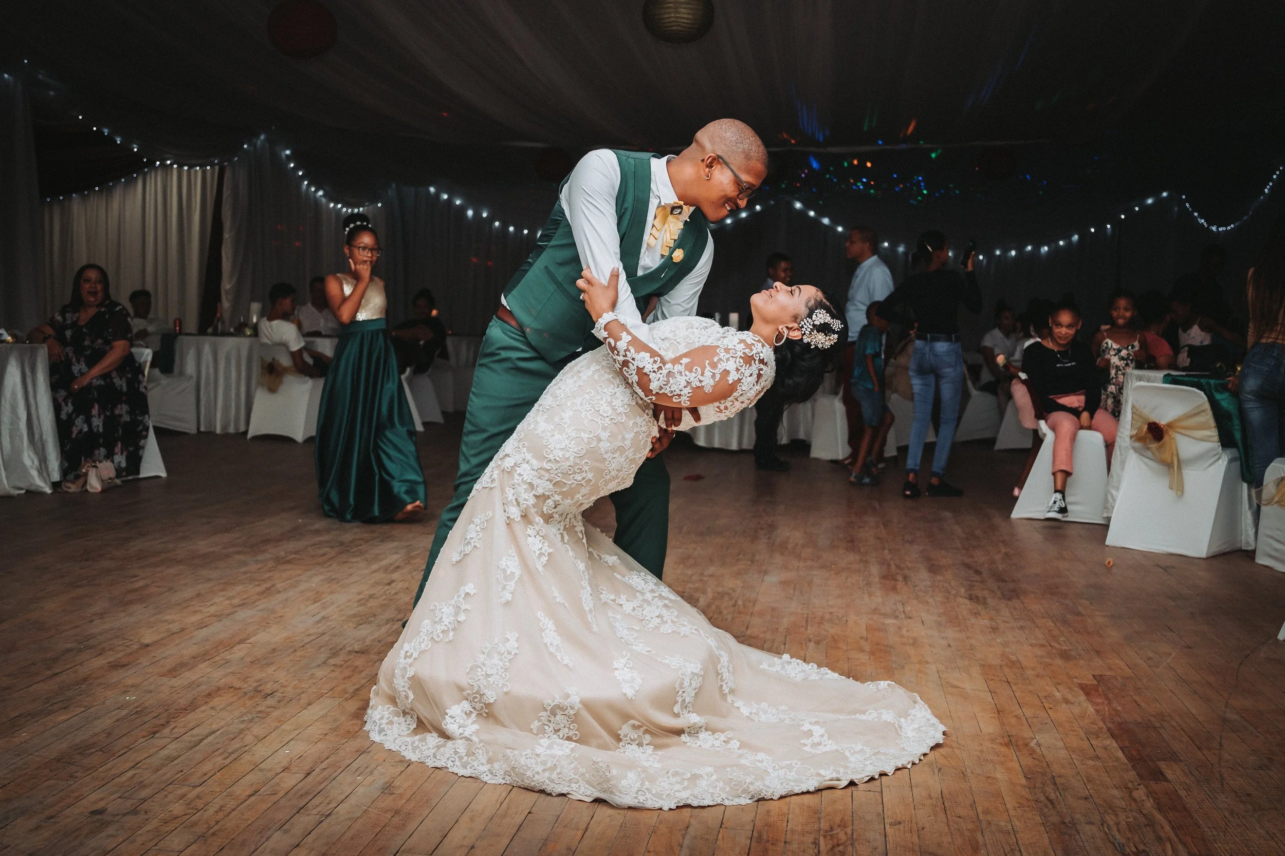 A bride and groom dancing at their wedding reception, with the groom bending over the bride who is looking up at him, smiling. The bride is in a white lace wedding dress, and the groom is in a green suit. Guests are visible in the background enjoying