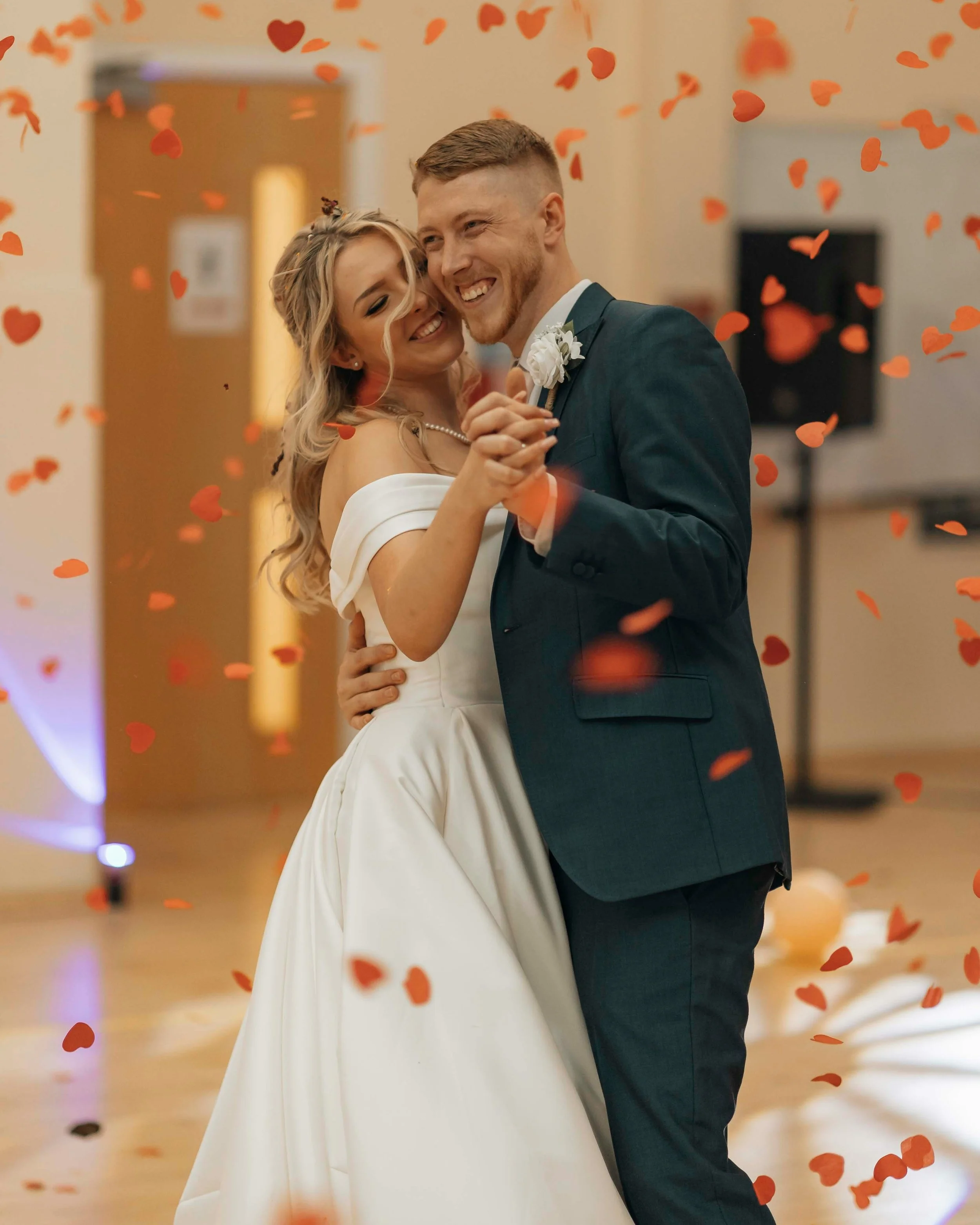 A bride and groom are dancing and smiling at their wedding reception, surrounded by floating red heart-shaped confetti.