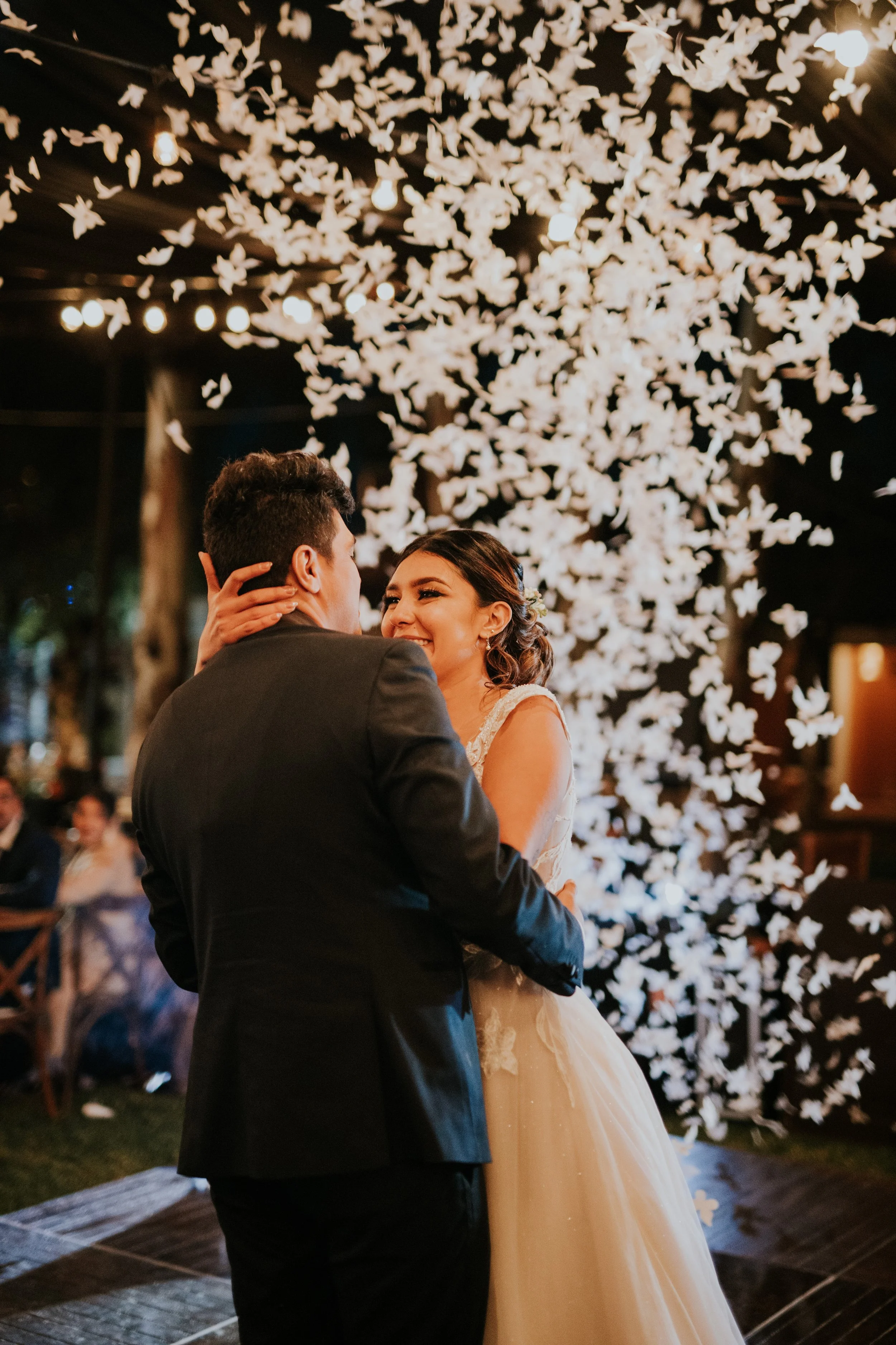 A newlywed couple sharing their first dance at their wedding reception, with a backdrop of white paper butterflies and warm lighting.