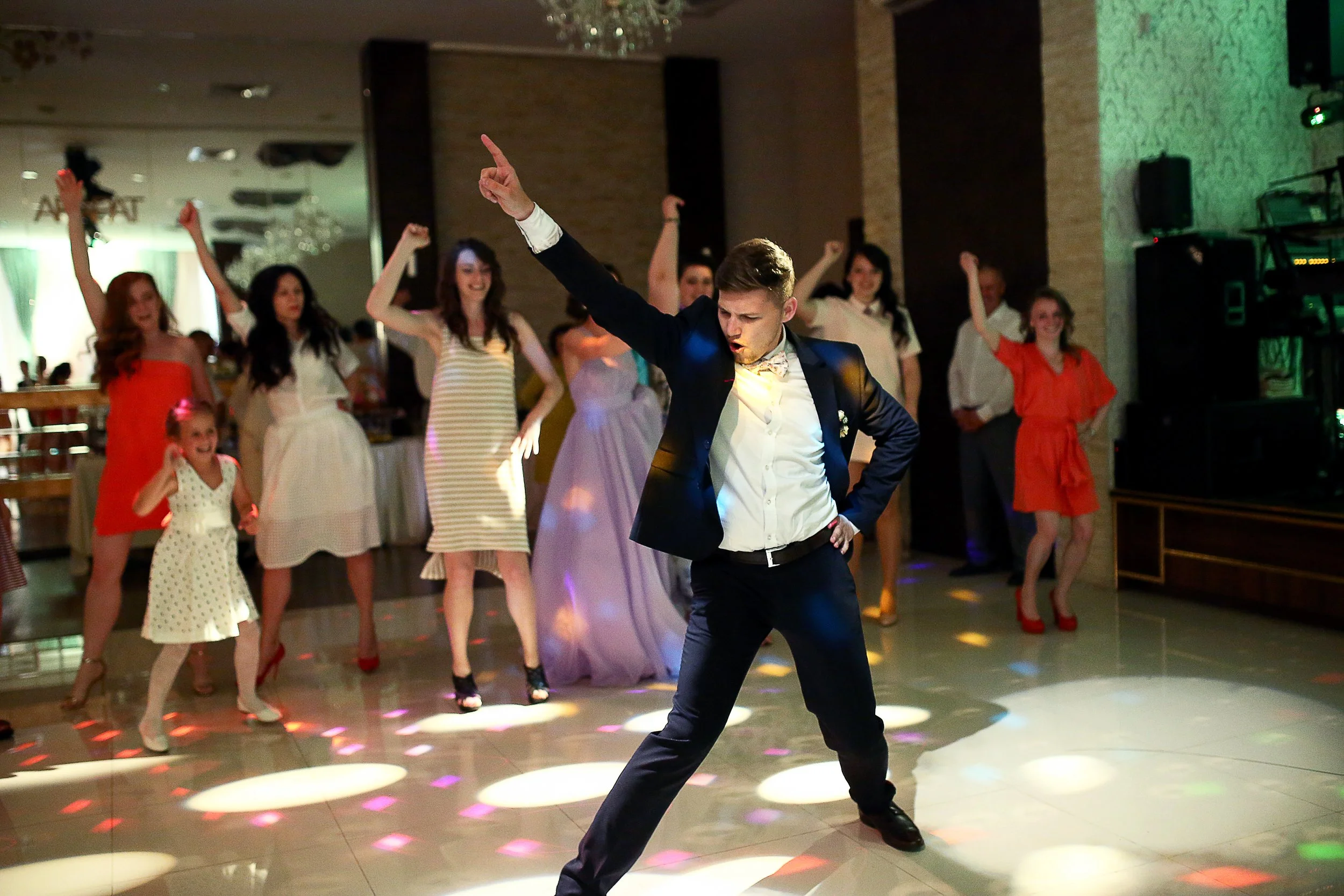 Young man in a suit dancing at a party with women and children watching and enjoying the performance, colorful lights on the dance floor.