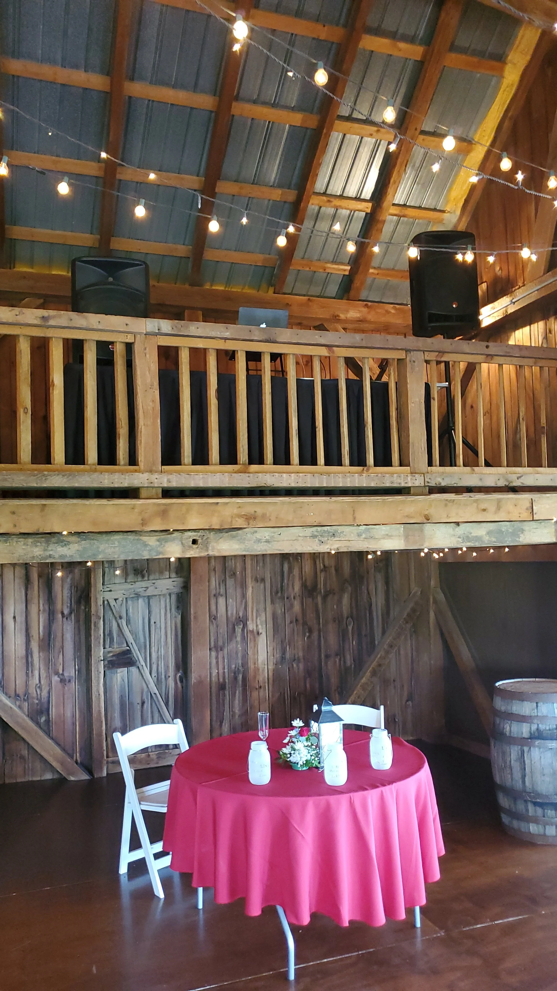 A rustic barn interior with a round table covered in a red tablecloth, set with four bottles of water, white folding chairs, and a floral centerpiece. The barn has wooden walls, a vaulted ceiling with exposed beams, string lights hanging, a balcony with speakers and a small DJ setup, and a wooden barrel in the corner.