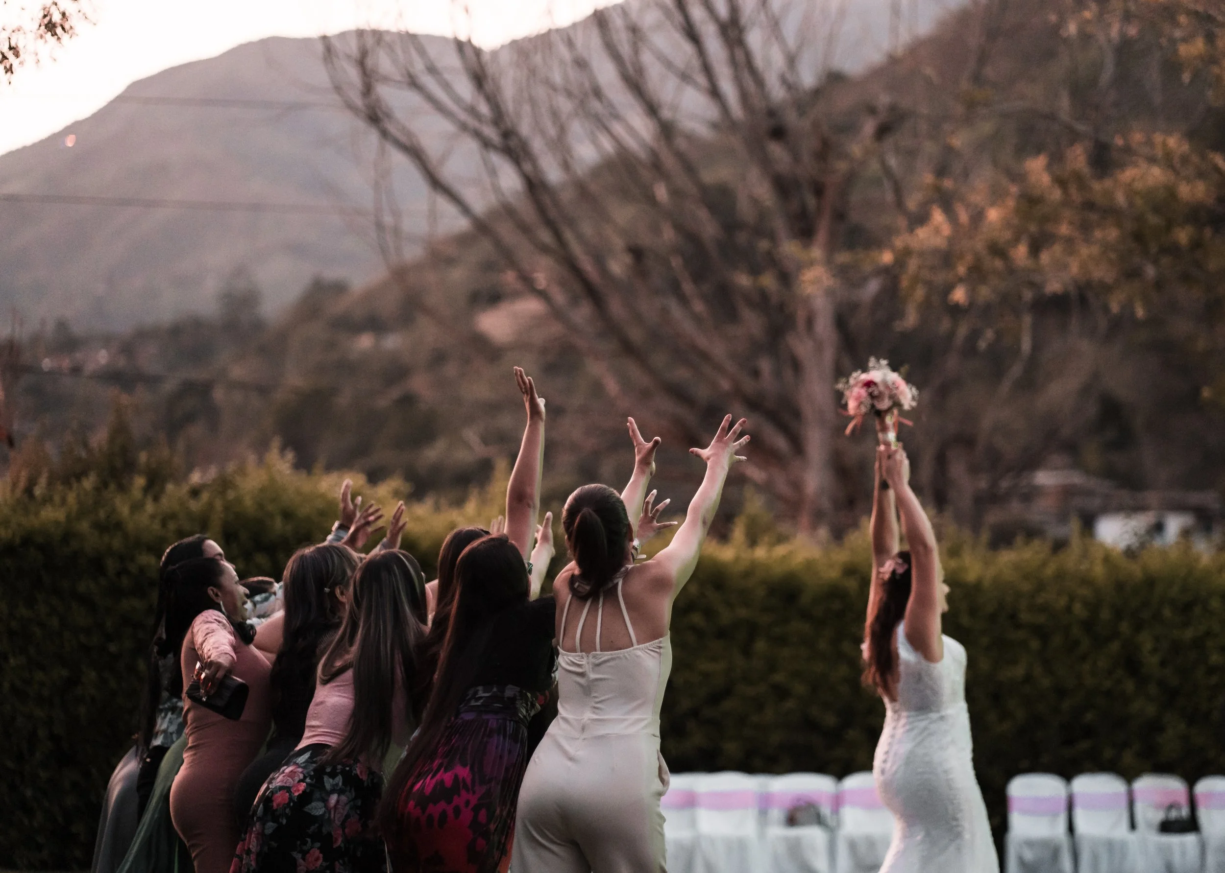 Group of women outdoors reaching towards a small bouquet of pink and white flowers raised in the air during sunset with mountains and trees in the background.