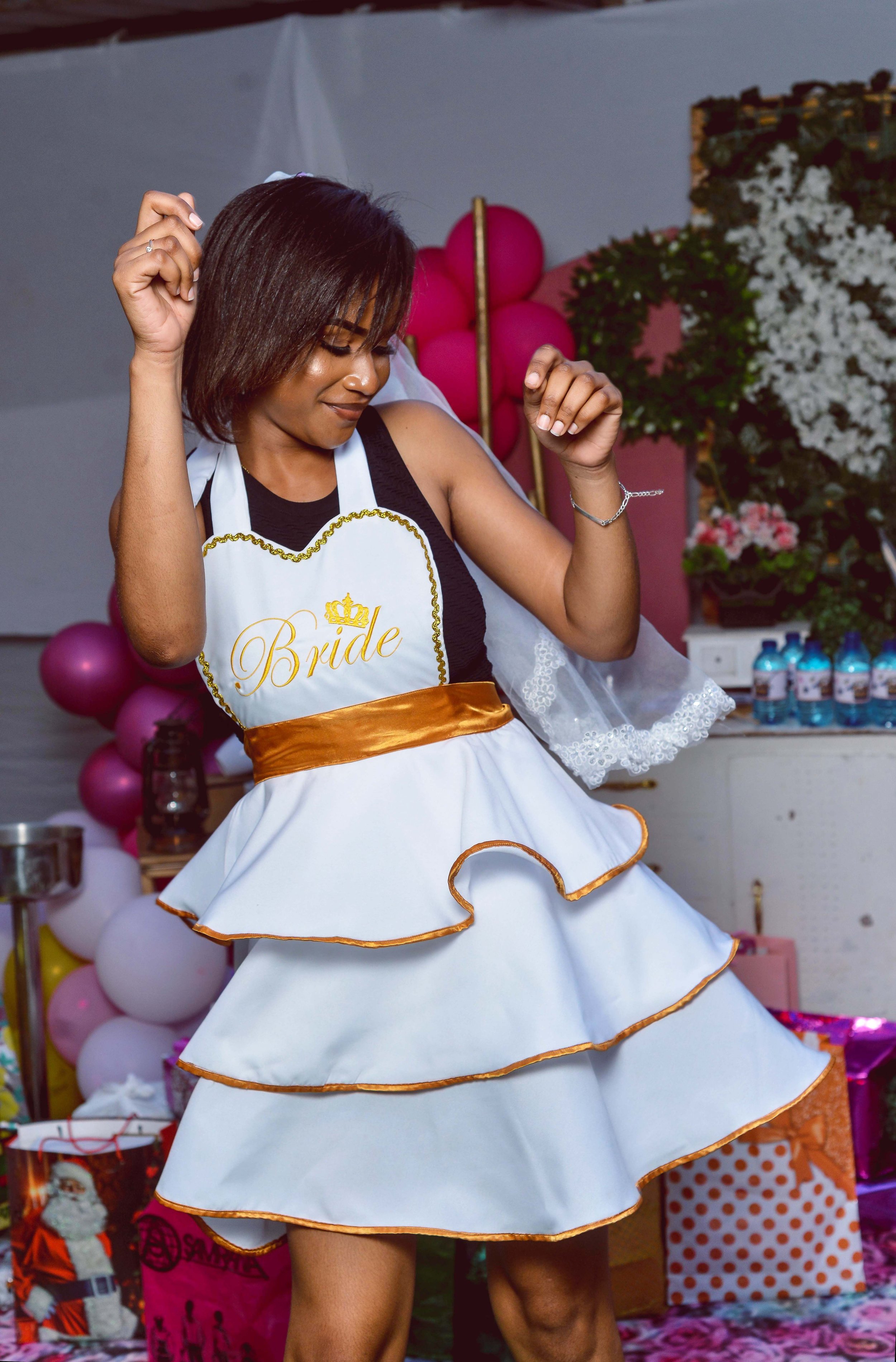 A woman at a celebration or party wearing a white dress with gold trim, a 'Bride' sash, and dancing. The background has pink and white balloons and floral decorations.