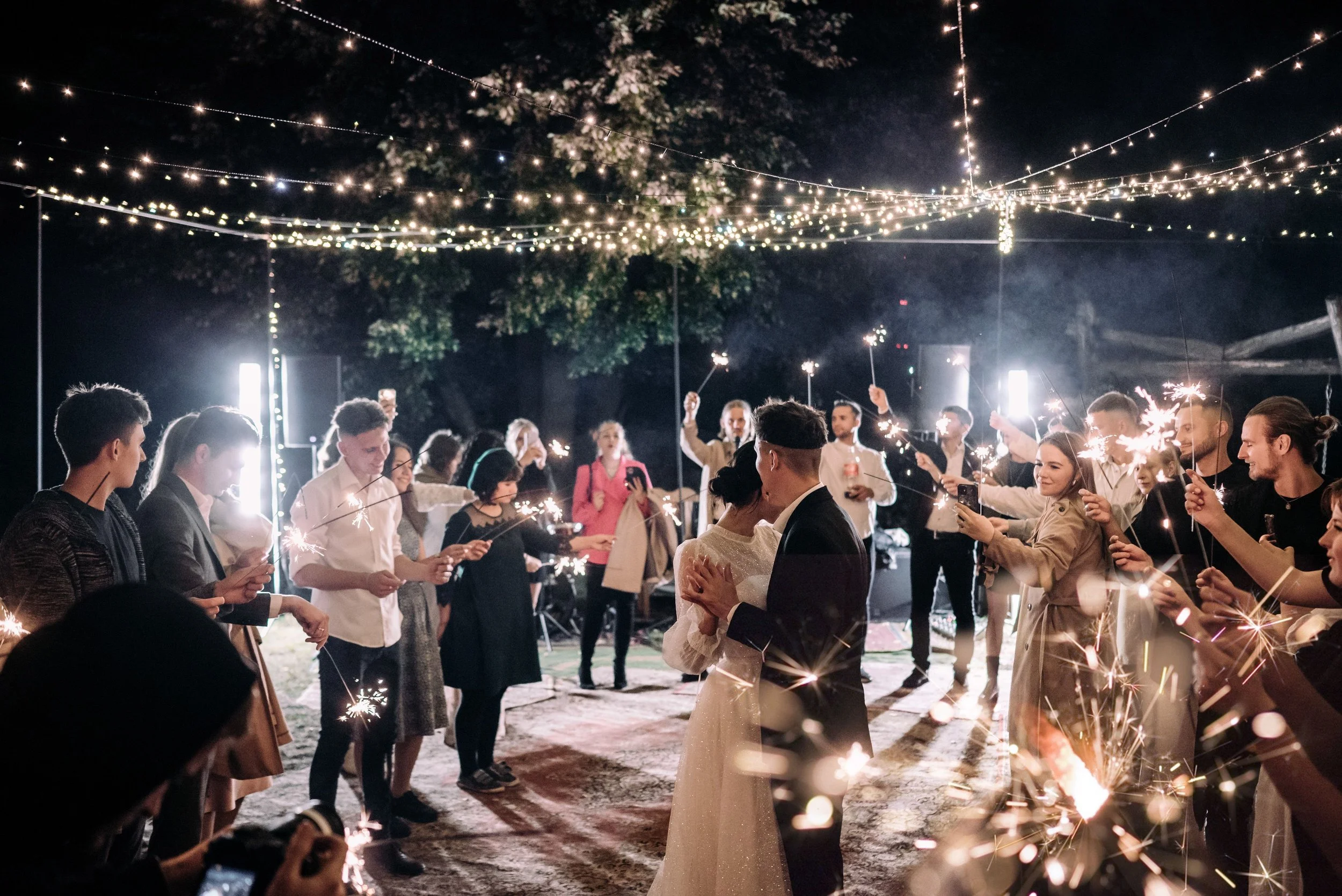 People celebrating at night with sparklers and string lights overhead, outdoor party scene.