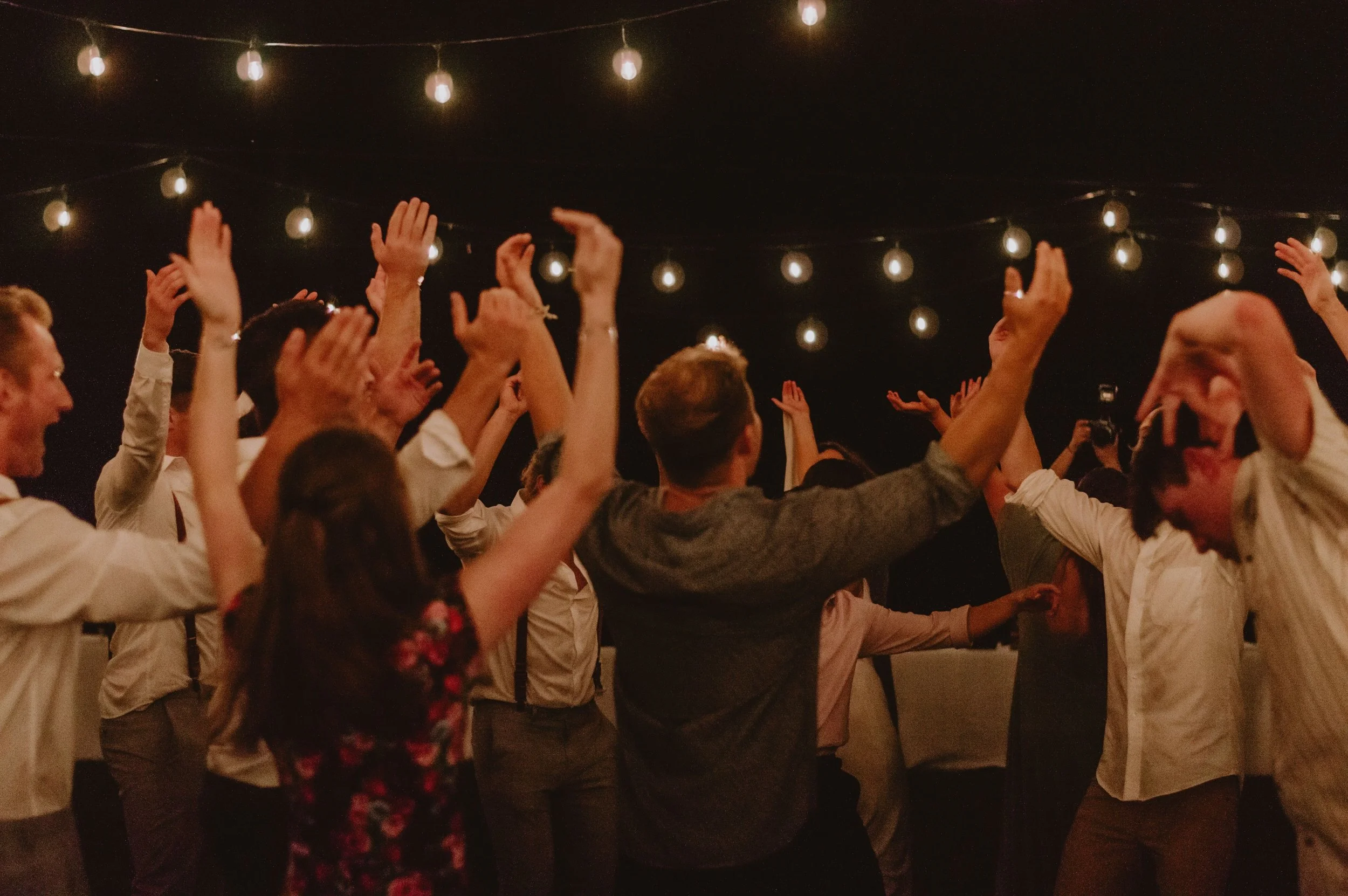 People dancing and raising their hands under string lights at an outdoor evening gathering.