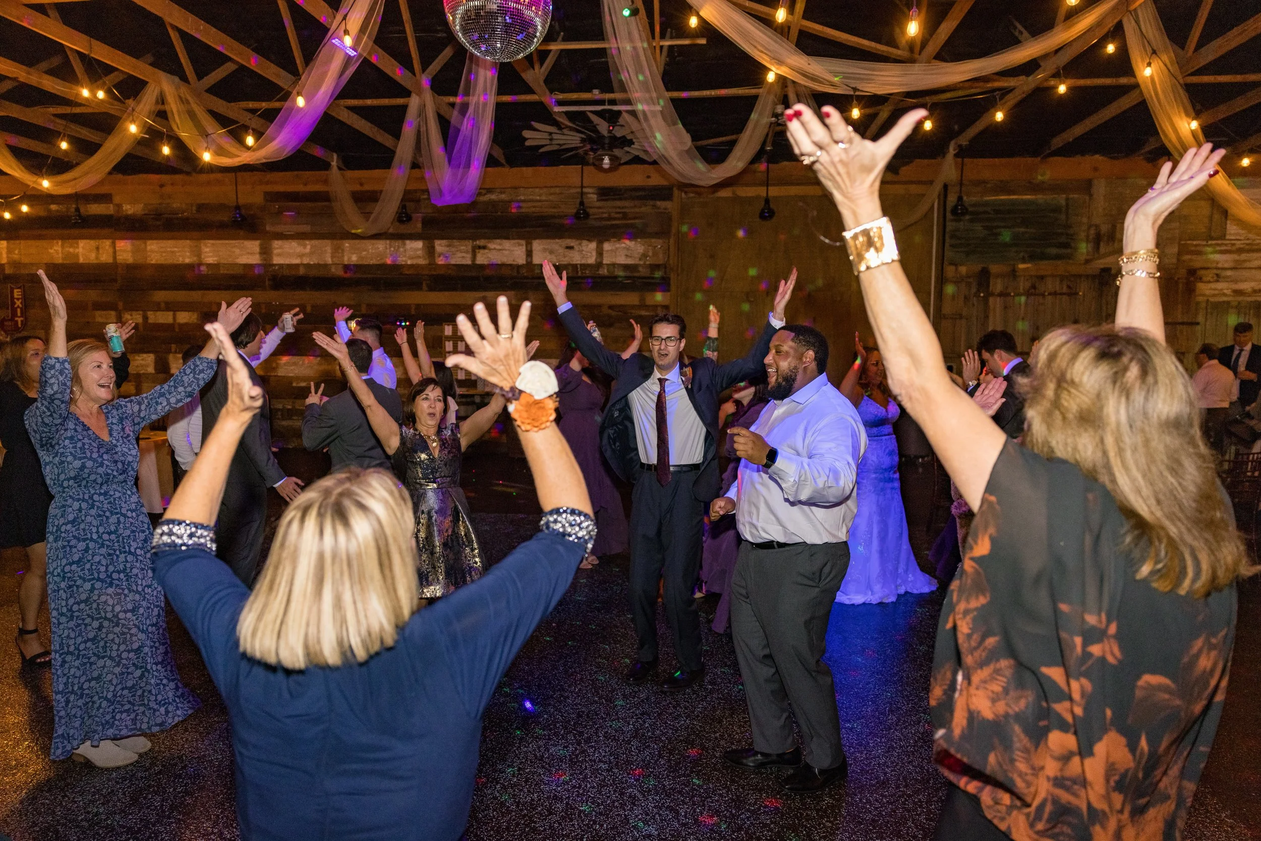People dancing and celebrating at a lively indoor party with a wooden interior, string lights, draped fabric, and a disco ball overhead.