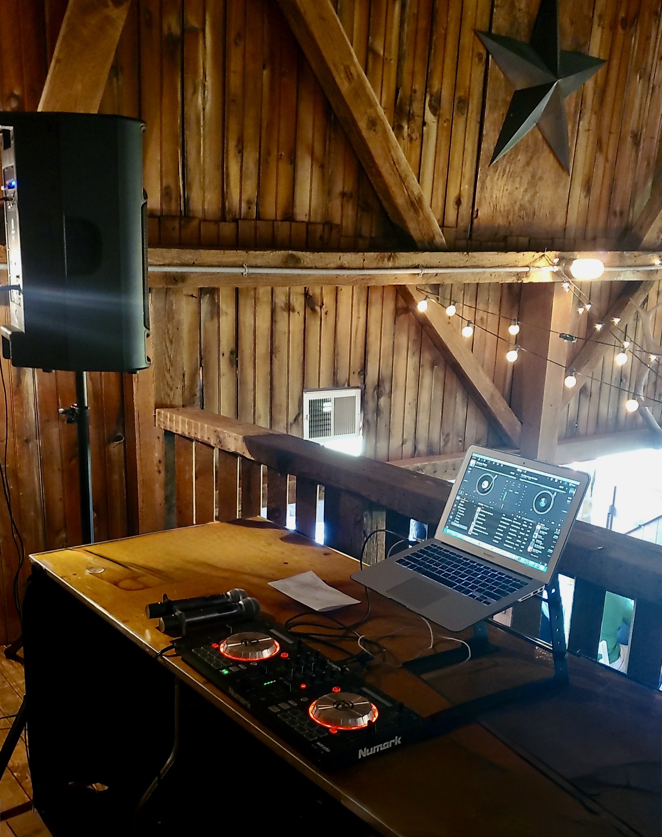 A DJ setup on a wooden table with a laptop, DJ controller, and headphones, in a rustic venue with wooden walls and string lights, and a large speaker to the left.