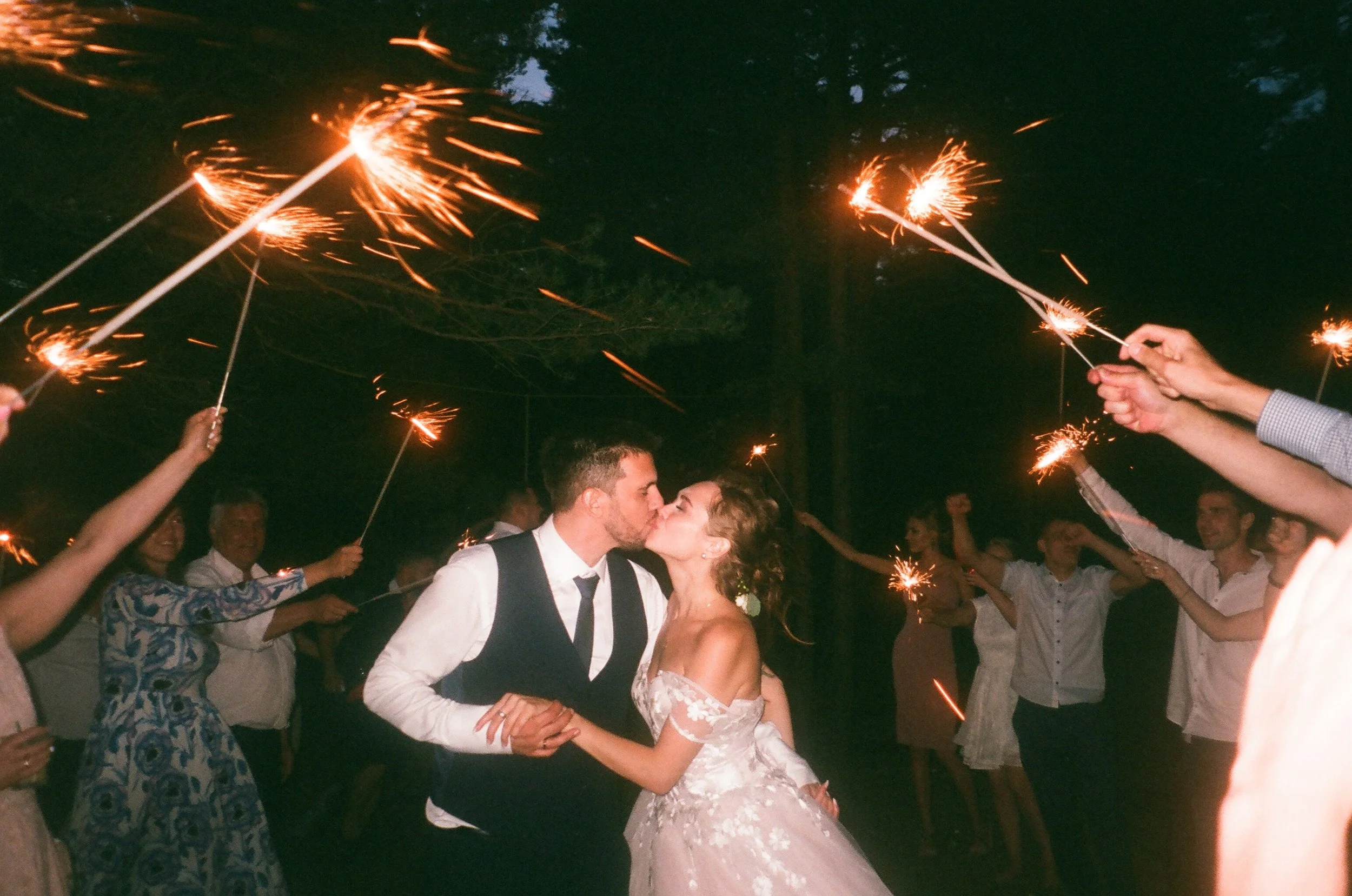 A bride and groom kiss amid guests holding sparklers during an outdoor night celebration.