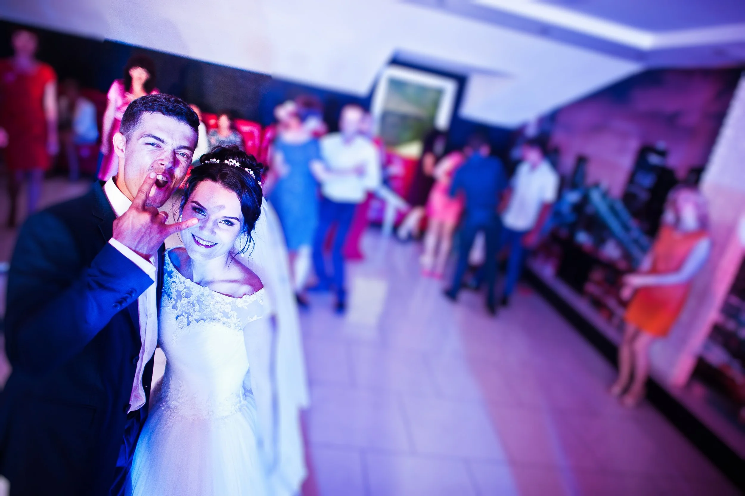 A newlywed couple taking a selfie at their wedding reception, with guests in the background.