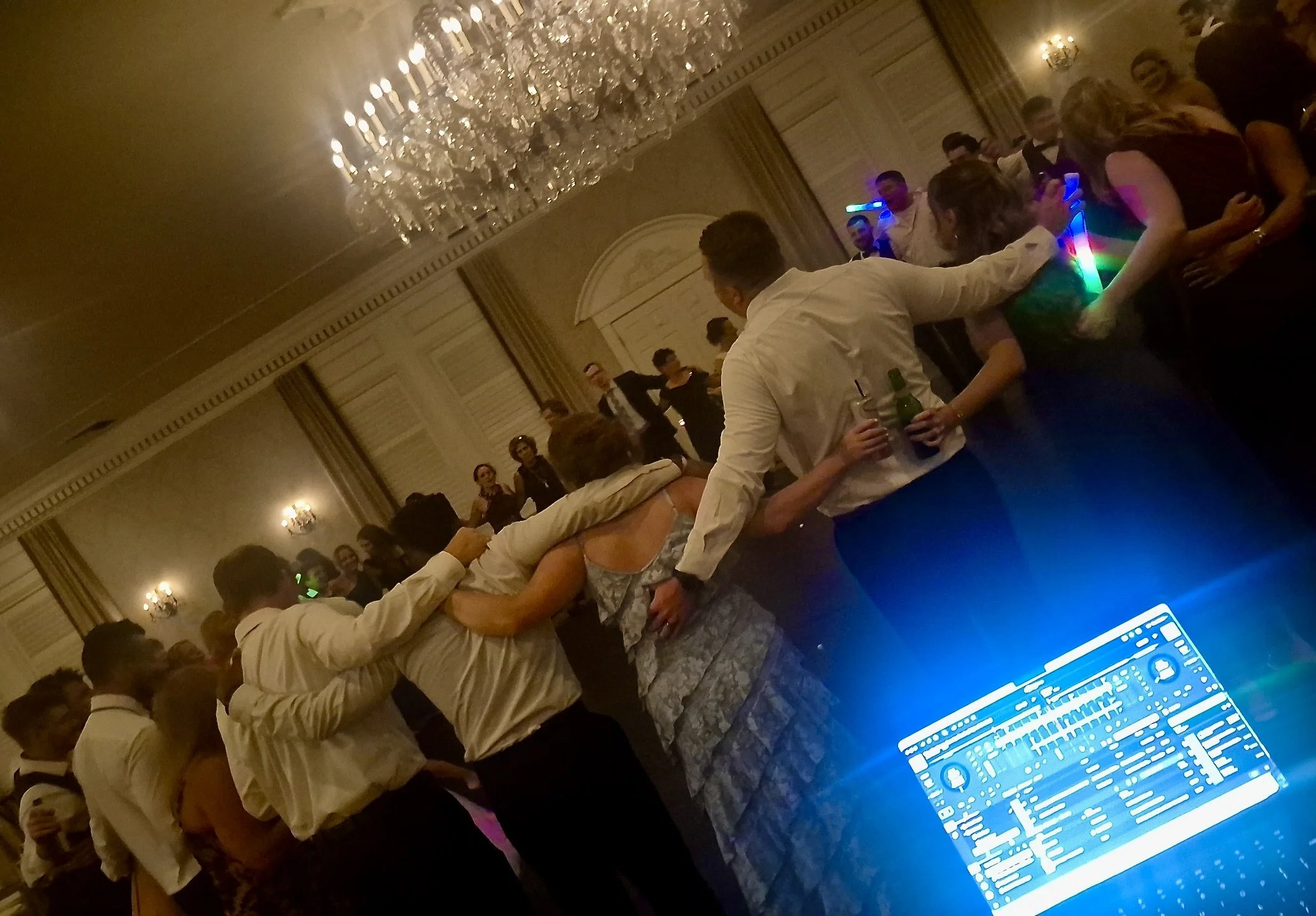 People dancing and socializing at a formal event in an elegant ballroom with ornate walls, chandeliers, and warm lighting, some holding drinks, and a DJ booth with a computer visible in the foreground.
