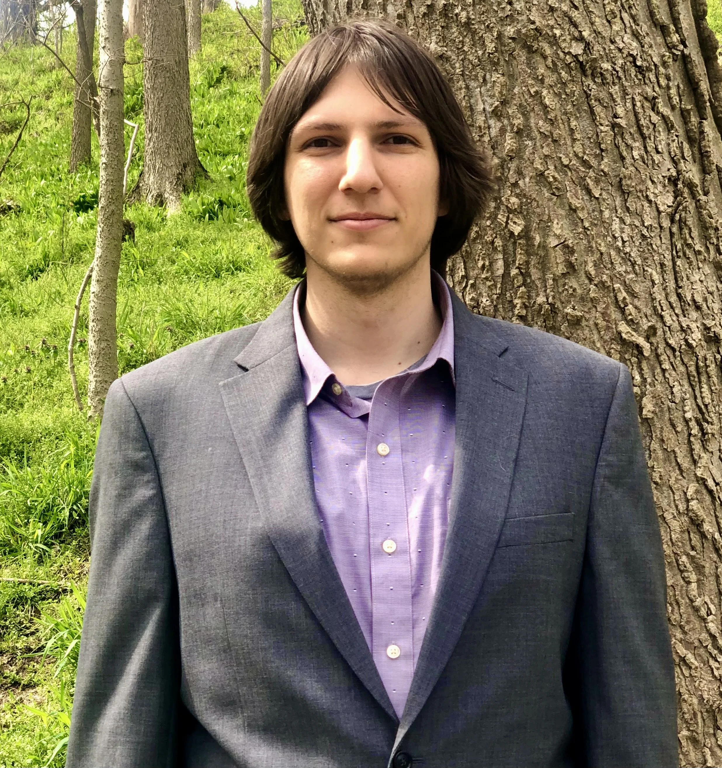 A young man with shoulder-length brown hair, wearing a gray suit jacket over a purple shirt, standing outdoors in front of a large tree with green grass and several smaller trees in the background.