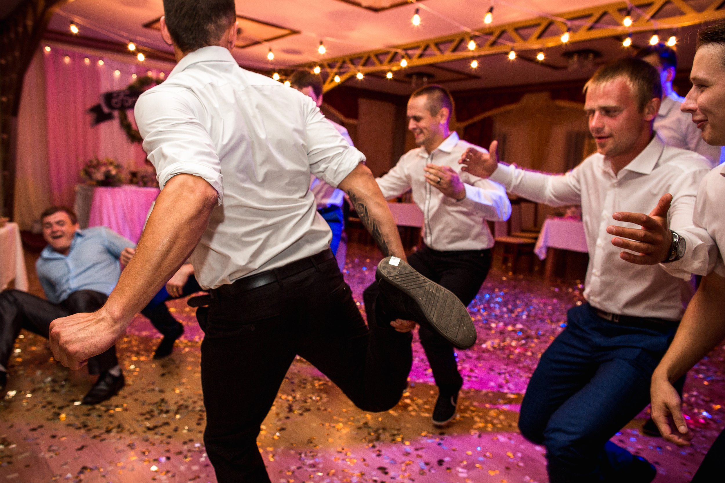 Group of men dancing and enjoying themselves at a lively indoor party with string lights and confetti on the floor.