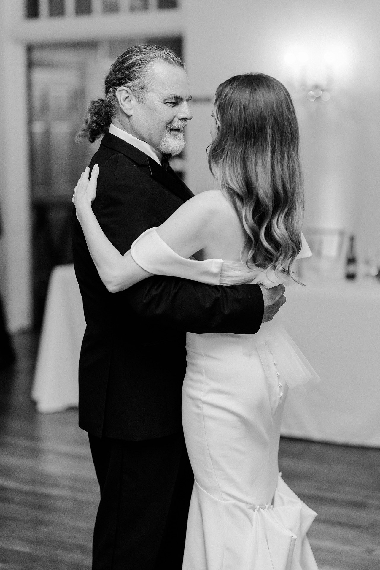 A black and white photo of a man and woman dancing together, likely at a wedding. The man has gray hair and a beard, wearing a dark suit, and the woman has long wavy hair, wearing a strapless white dress. They are smiling and gazing at each other.
