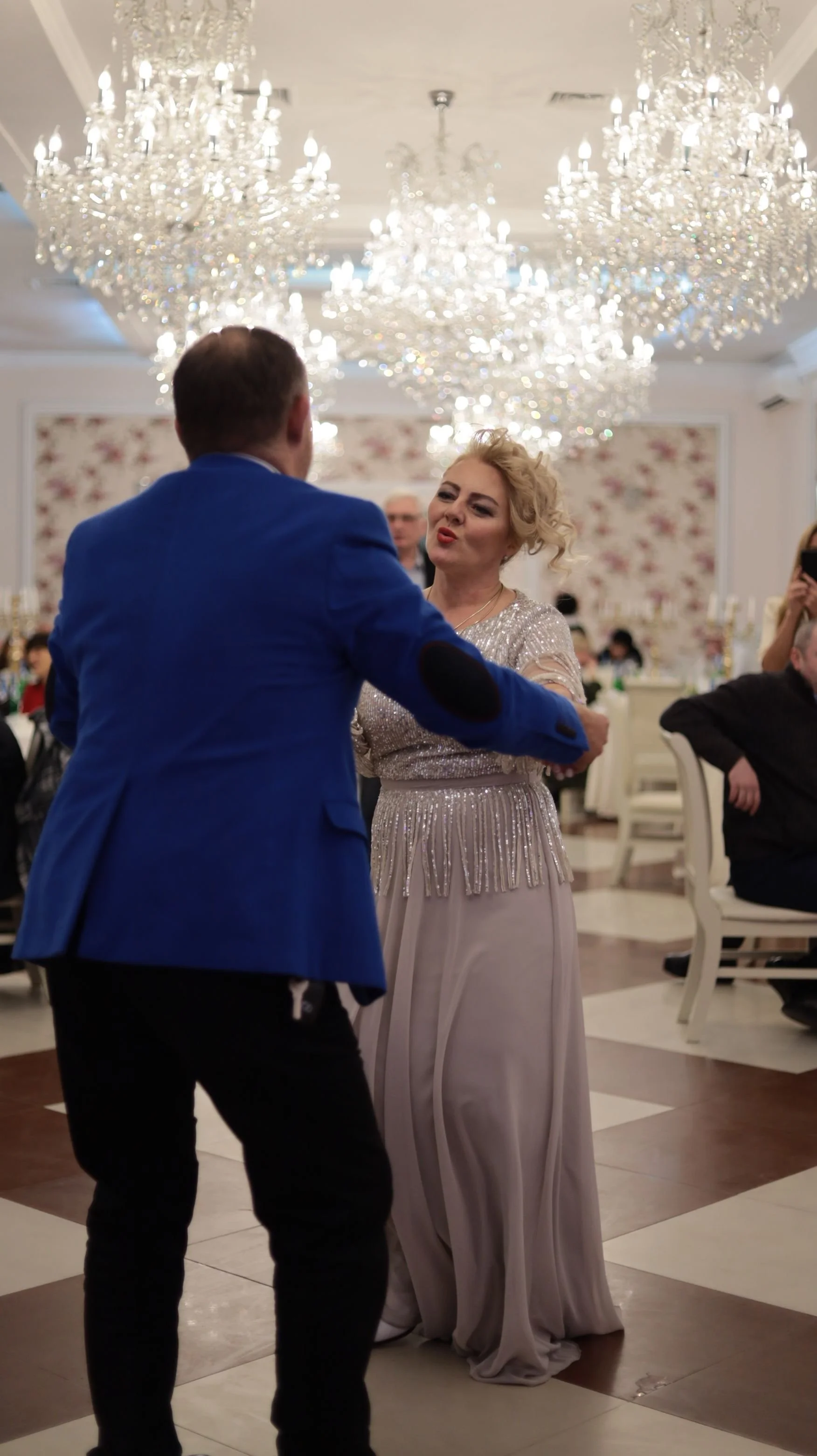 A man in a blue jacket and black pants dancing with a woman in a long, sparkling silver gown in a decorated reception hall with chandeliers.