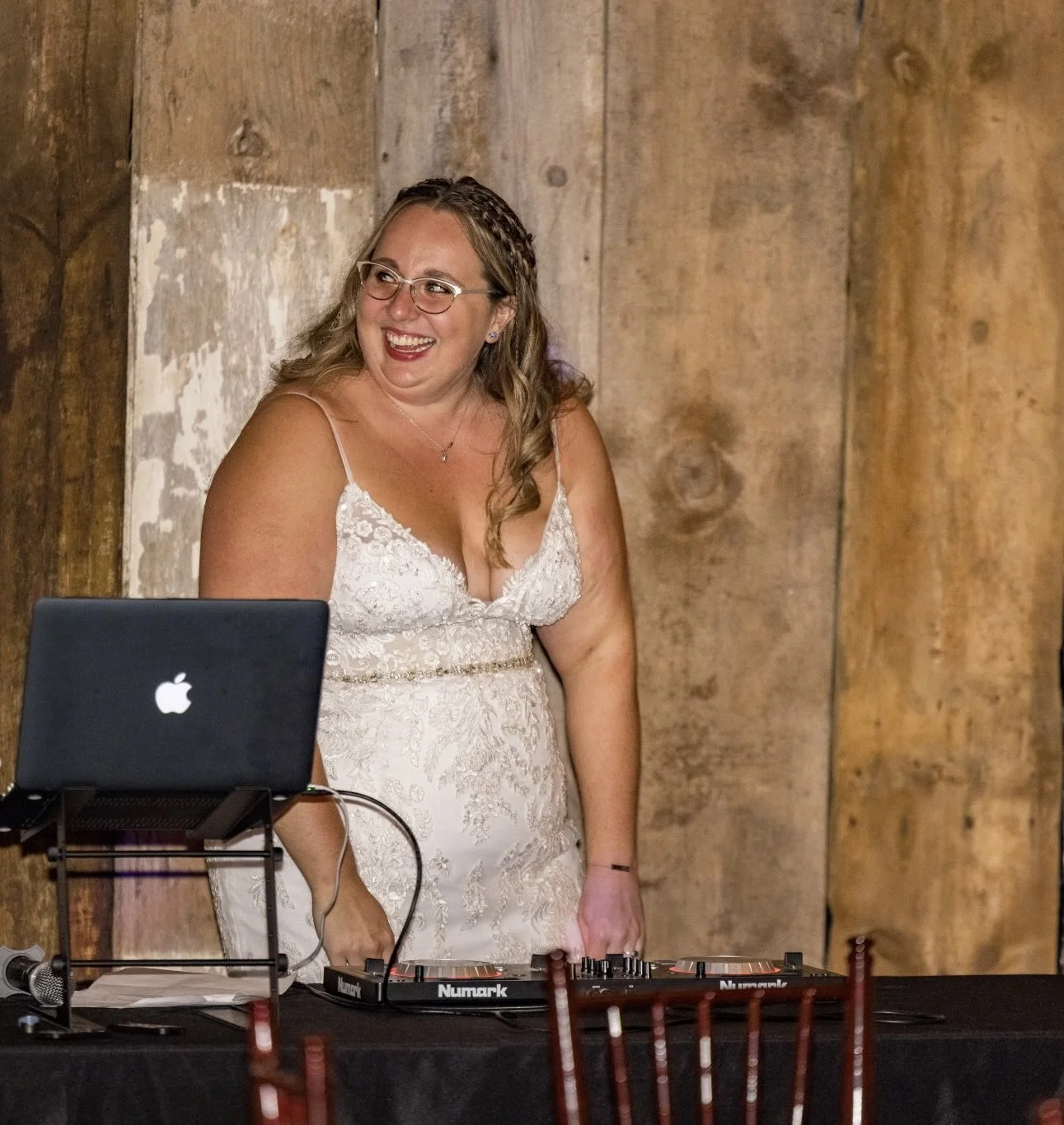 A woman in a wedding dress, smiling and standing behind DJ equipment, with a laptop on a stand in front of her, in front of a rustic wooden wall.