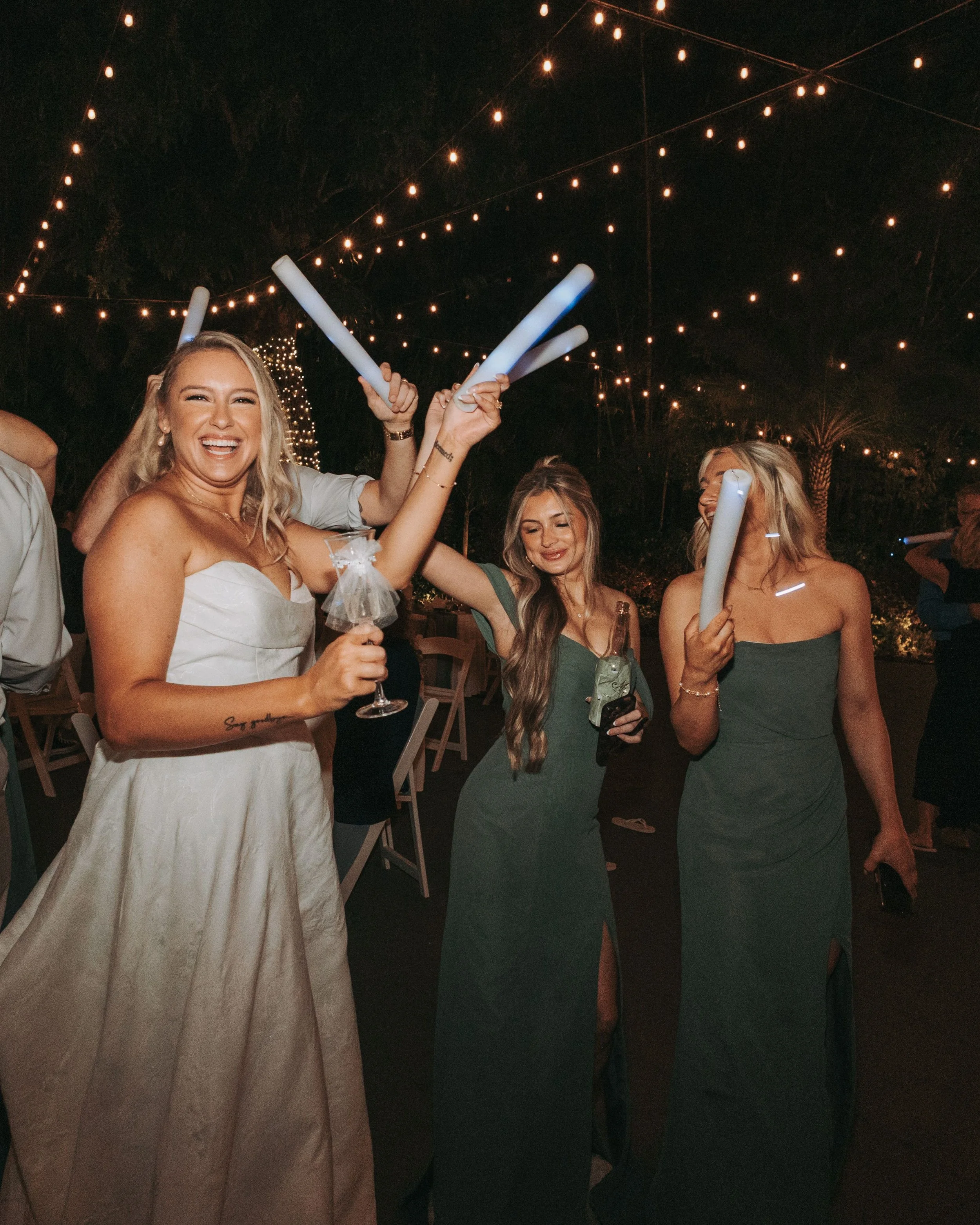 Nighttime outdoor celebration with string lights, women in formal dresses holding glow sticks, smiling, and enjoying each other's company.