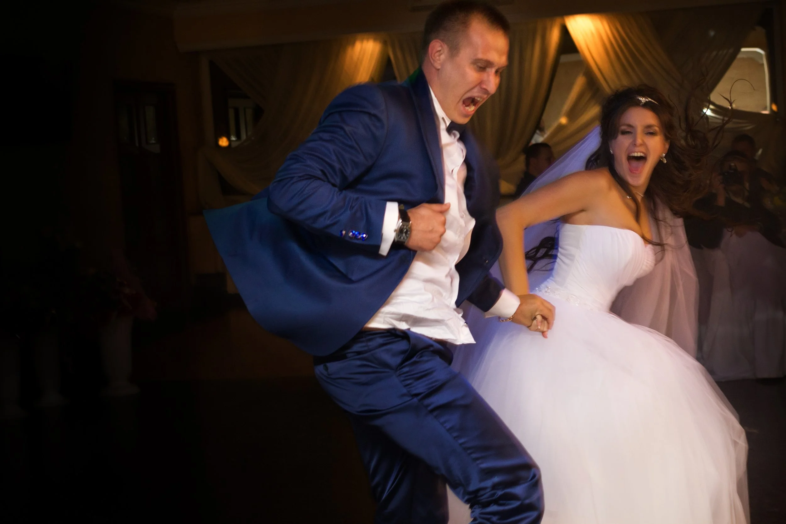 A man and woman dancing at a wedding reception. The man is wearing a blue suit and white shirt, and the woman is wearing a white wedding gown. Both are smiling and appear to be having fun.