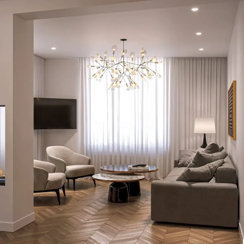 Modern living room with beige armchairs, a gray sofa with pillows, a marble-top coffee table with magazines, a wall-mounted flat-screen TV, a floor lamp, white curtains, a unique gold chandelier, and wooden herringbone flooring.