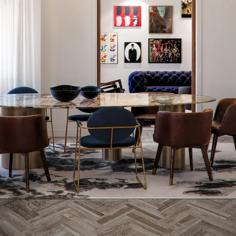 A dining room with a marble-top table and six chairs, three upholstered brown chairs, one navy blue chair with gold legs, and two matching dark blue chairs. The floor has a patterned rug and herringbone wood flooring. The wall in the background has seven pieces of modern and abstract artwork, and a purple tufted velvet sofa is visible through an opening in the wall.