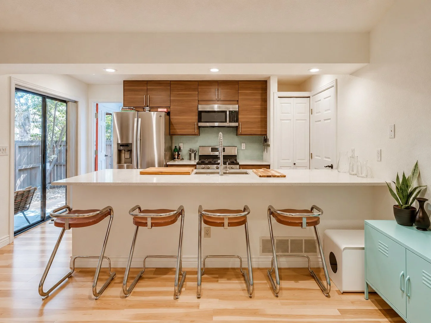 Modern kitchen with wooden cabinets, stainless steel appliances, white countertops, and four brown barstools at the breakfast bar, with sliding glass door leading outside