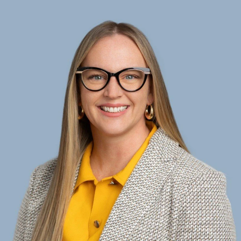 A woman with long blonde hair wearing black glasses, a yellow blouse, and a patterned blazer, smiling against a light blue background.