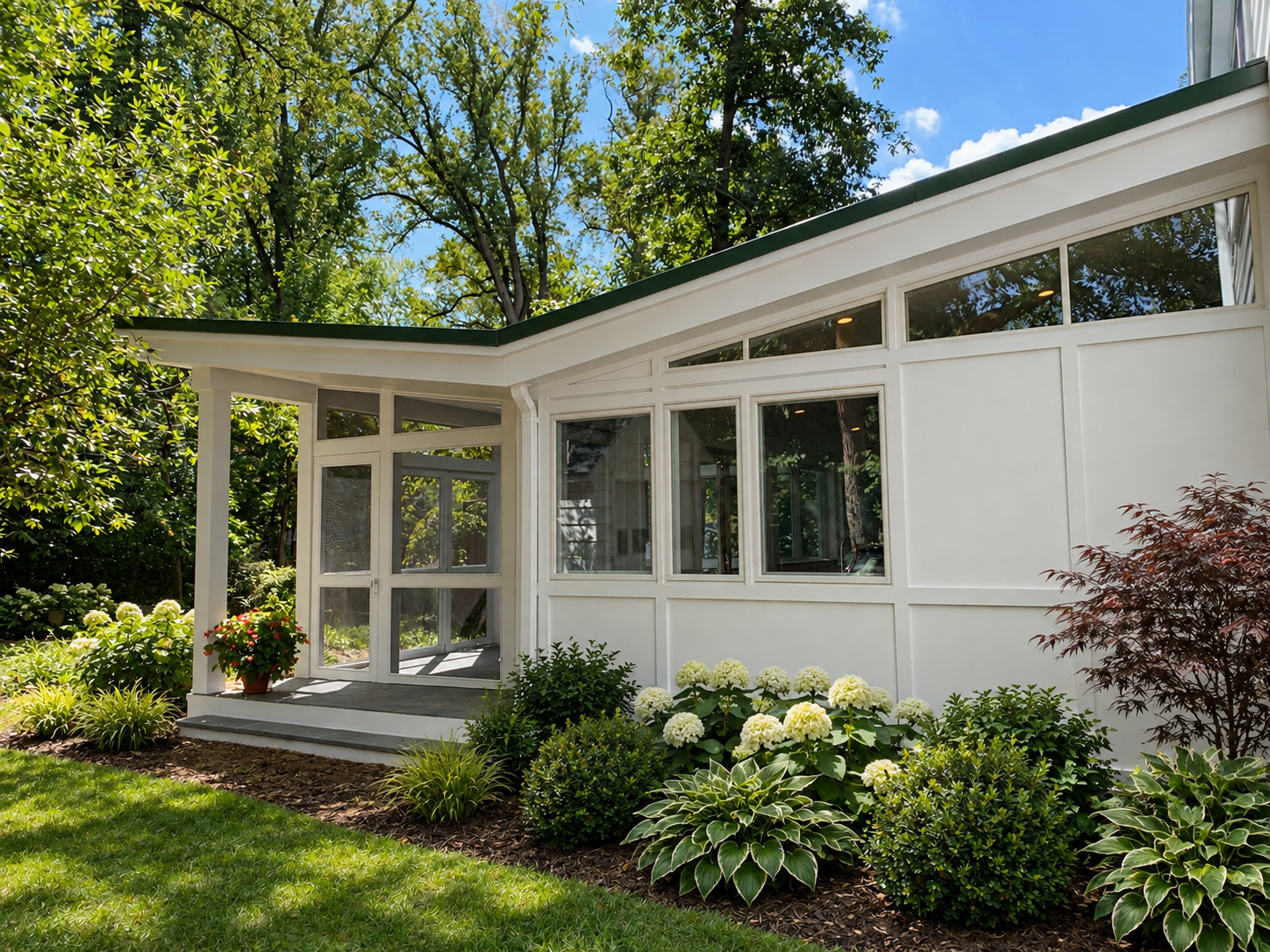 A modern glass sunroom attached to a white house, surrounded by lush green trees, shrubs, and blooming flowers, with a small patio area.