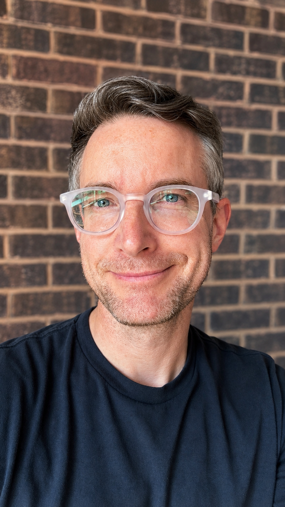 A smiling man with short brown hair, wearing clear glasses and a navy t-shirt, standing in front of a brick wall.