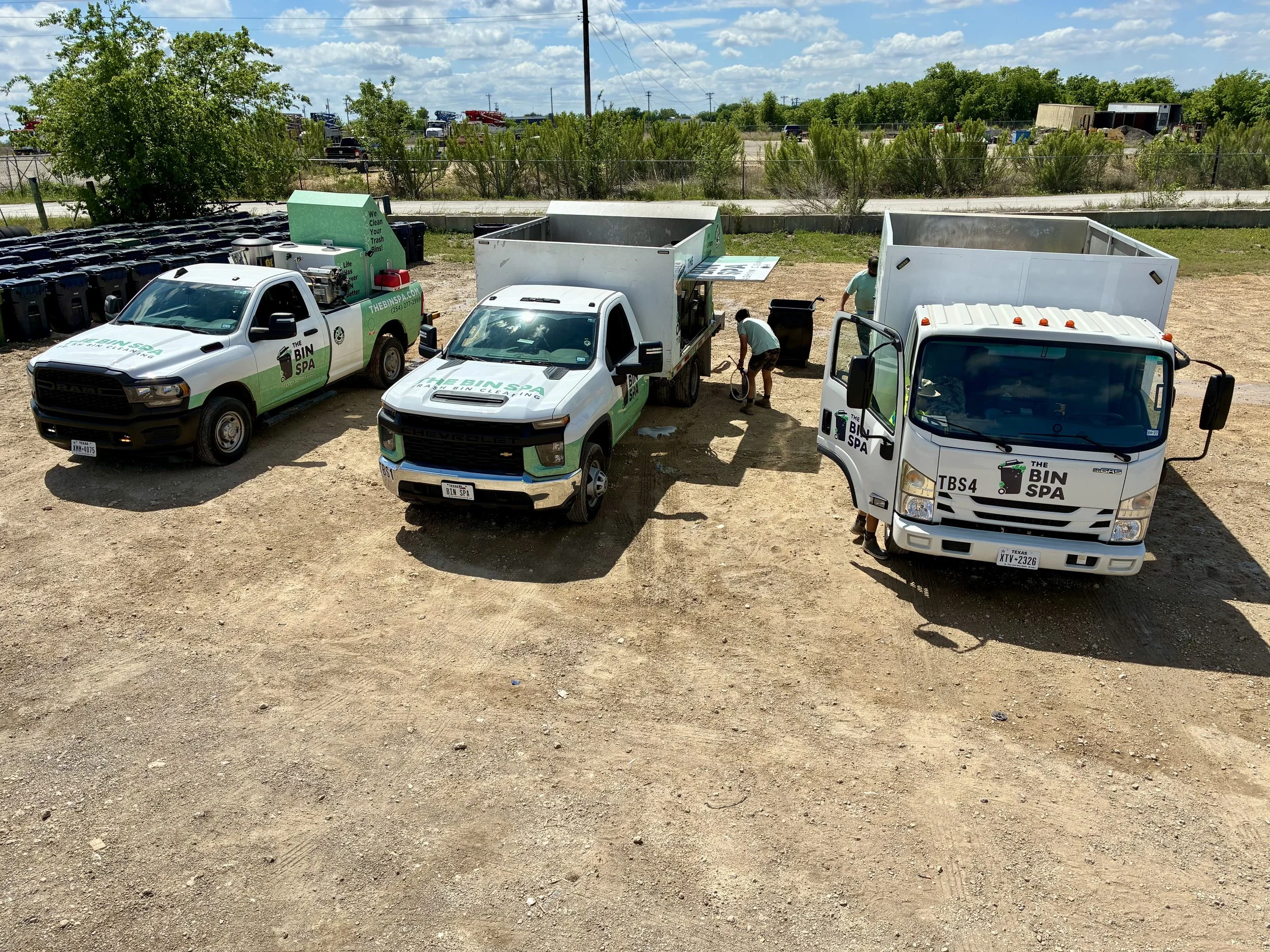 Three work trucks with branding for 'The Bin Spa' are parked on a dirt lot, with two workers loading trash into large open-top containers. Behind the trucks, there's a chain-link fence and a background of trees, bushes, and a partly cloudy sky.
