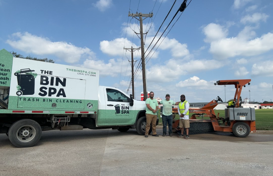 Four men standing next to a trash bin cleaning truck and a street sweeper on a road, with utility poles and a blue sky with clouds in the background.