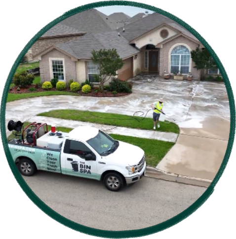 A cleaning professional power washing a house driveway while a service truck is parked nearby in a residential neighborhood.