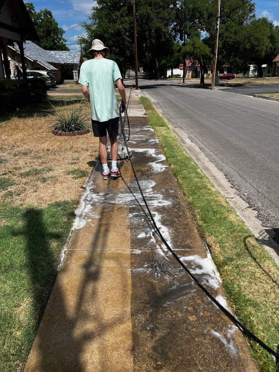 Person wearing a light green shirt, black shorts, and white shoes washing the sidewalk with a hose, creating soap bubbles on the concrete.