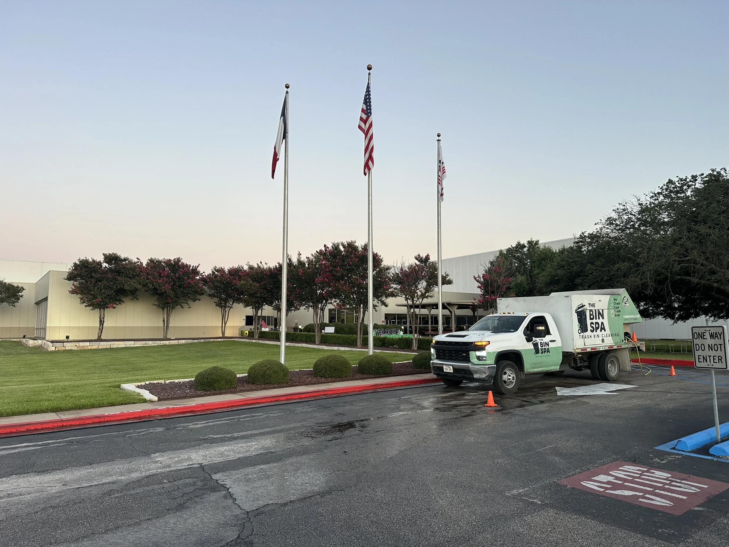 Three flagpoles with American flags, a small trash bin cleaning truck labeled 'The Bin Spa,' and a sign that says 'One Way Do Not Enter' in front of a building surrounded by trees and bushes with pink flowers.