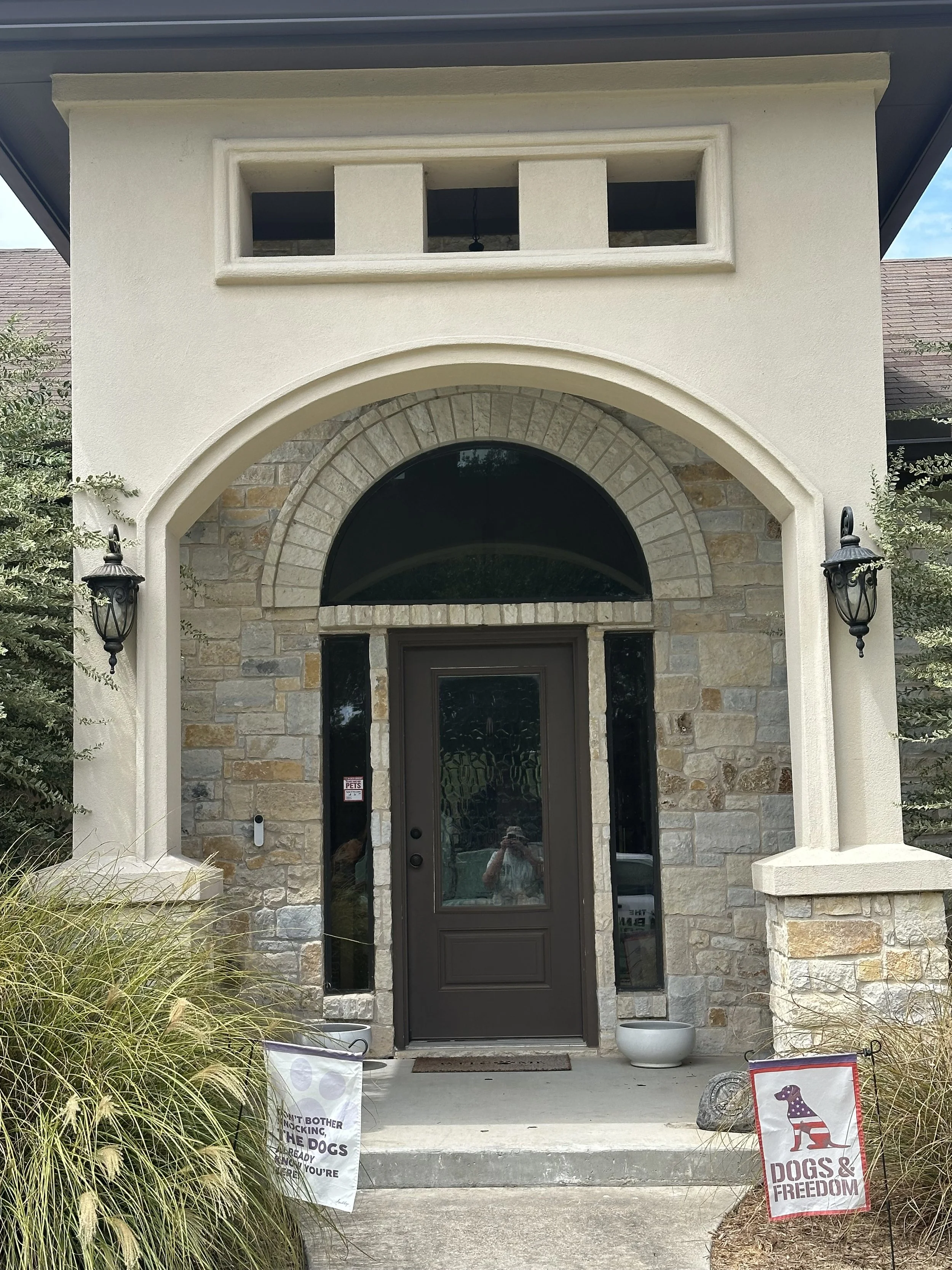 The image shows the front porch of a house with a stone and stucco facade, a black front door, and a decorative arched window above the door. Two black lantern-style wall sconces are mounted on either side of the door. There are signs placed on the ground near the steps that read 'DOGS & FREEDOM' and another with pet-related messages. The porch is surrounded by plants and decorative items.