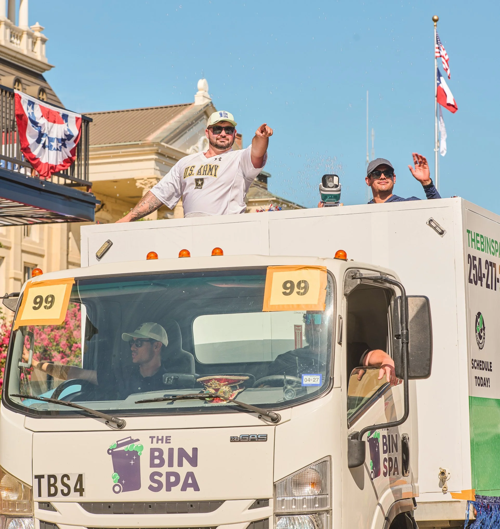 A white truck with colorful signage reading "The Bin Spa" and phone number, decorated with banners and American flags, carrying people celebrating, one in U.S. Army attire, during a parade with a clear blue sky background.