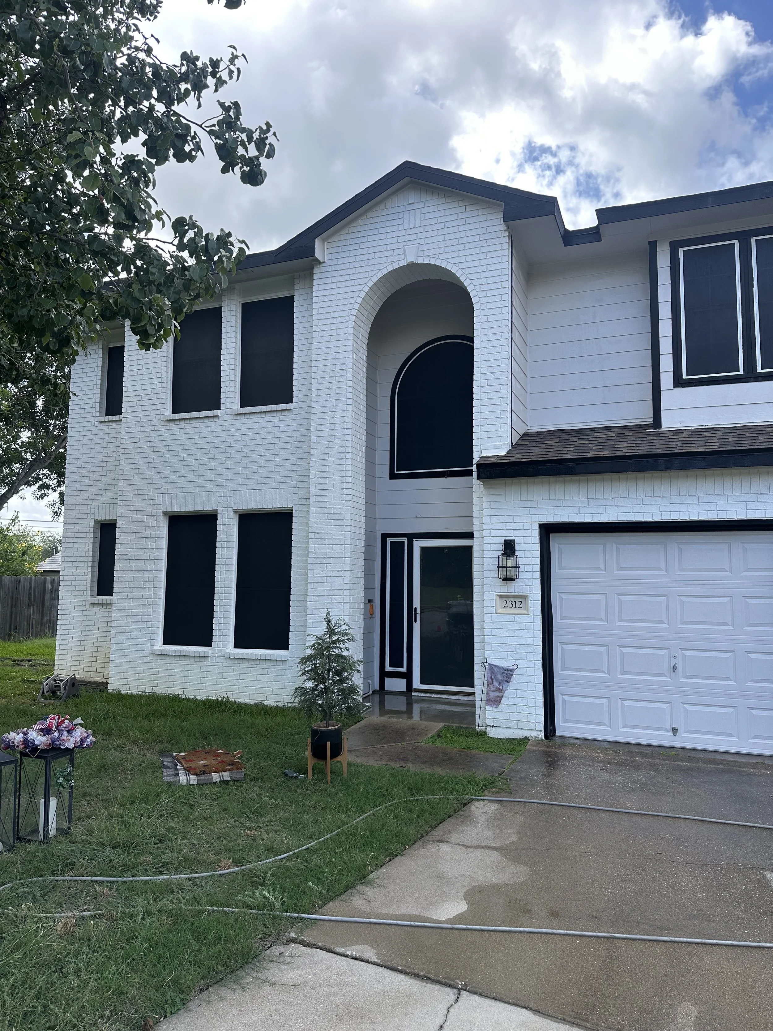 A new white two-story house with black accents and a three-car garage, surrounded by a green lawn with a small tree in front and a flower bed. The sky is partly cloudy.