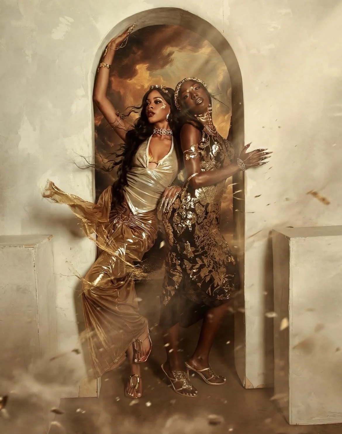 Two women in gold and black ornate dresses posing in front of a painted background with stormy clouds, surrounded by dust and debris.