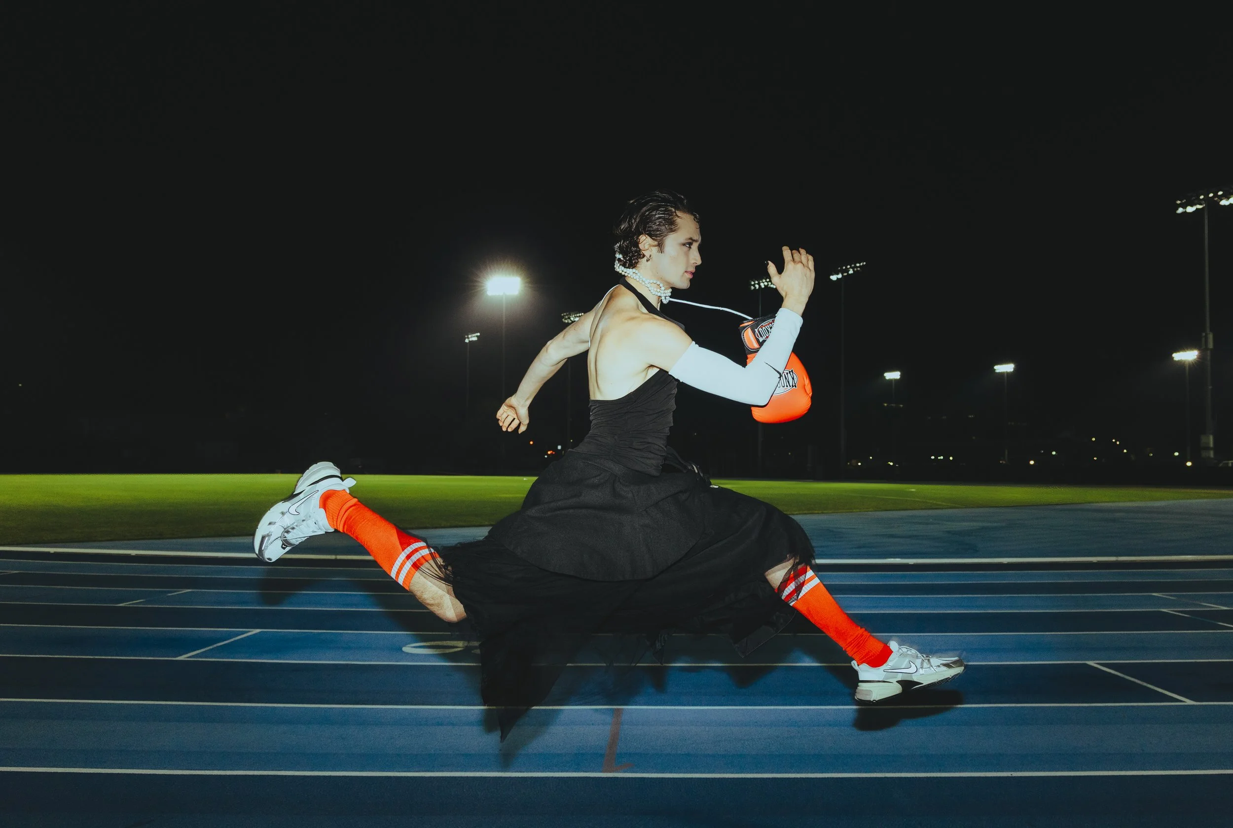 A person stretching on a running track at night, wearing athletic gear, with stadium lights illuminating the field.