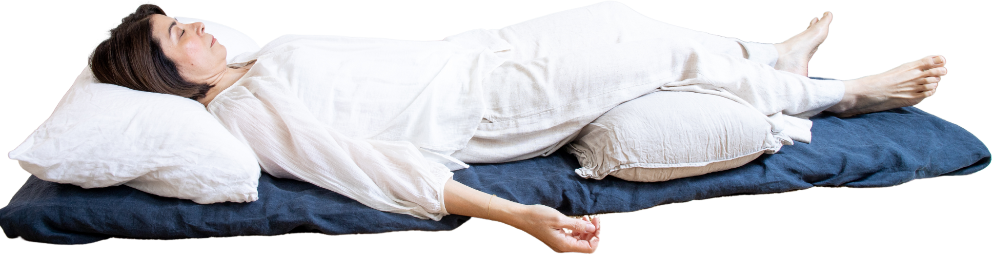 Una mujer acostada de lado en posición de yoga o meditación, en ropa cómoda, sobre un colchón con varias almohadas.
