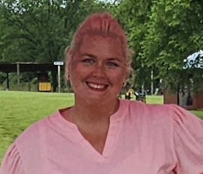 A woman with light skin, red hair styled up, smiling outdoors in a park with trees and grass, wearing a pink top.