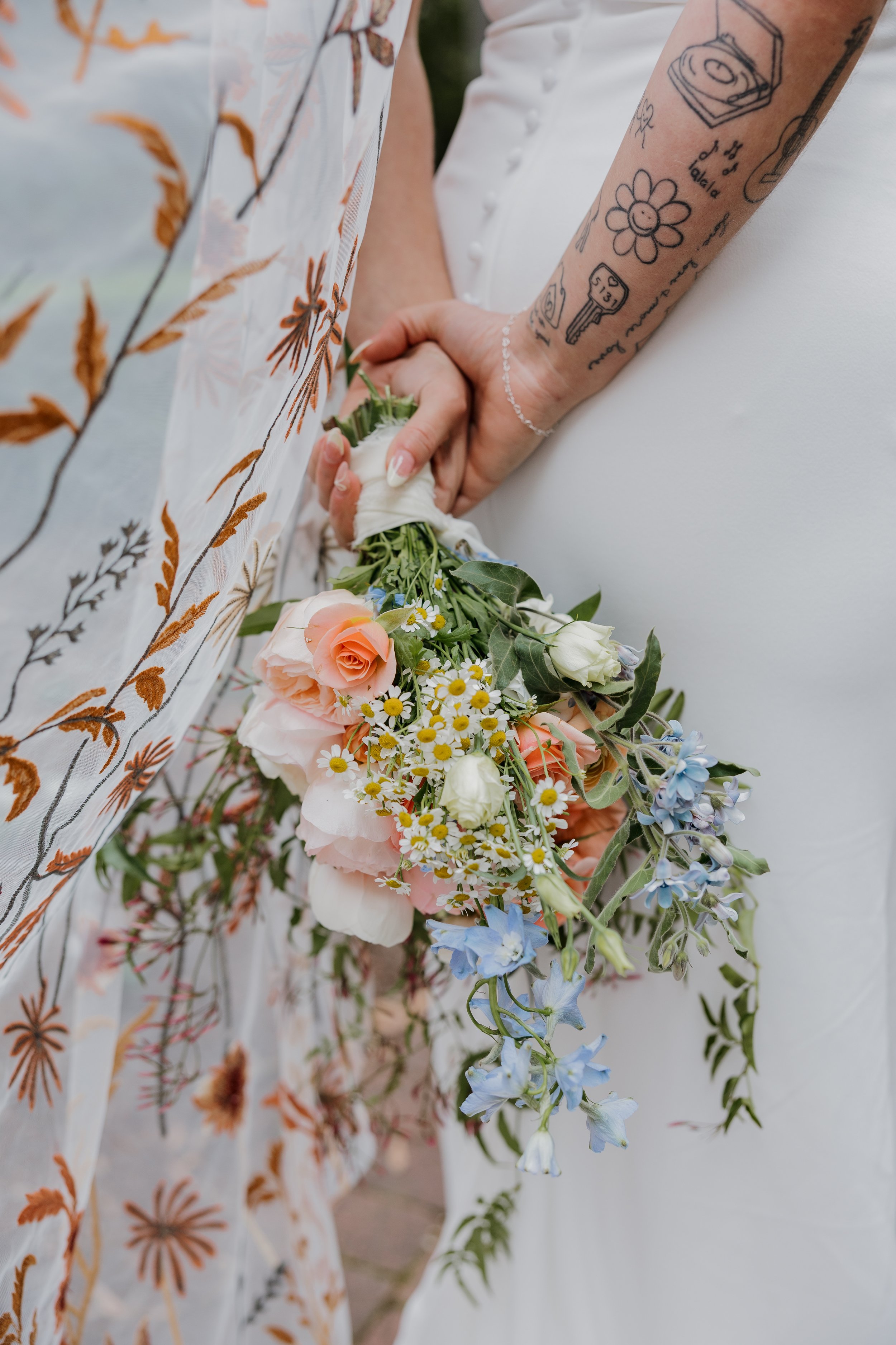 A couple holding hands with the woman holding a bouquet of flowers, featuring peach roses, white and yellow daisies, and blue flowers, during a wedding.