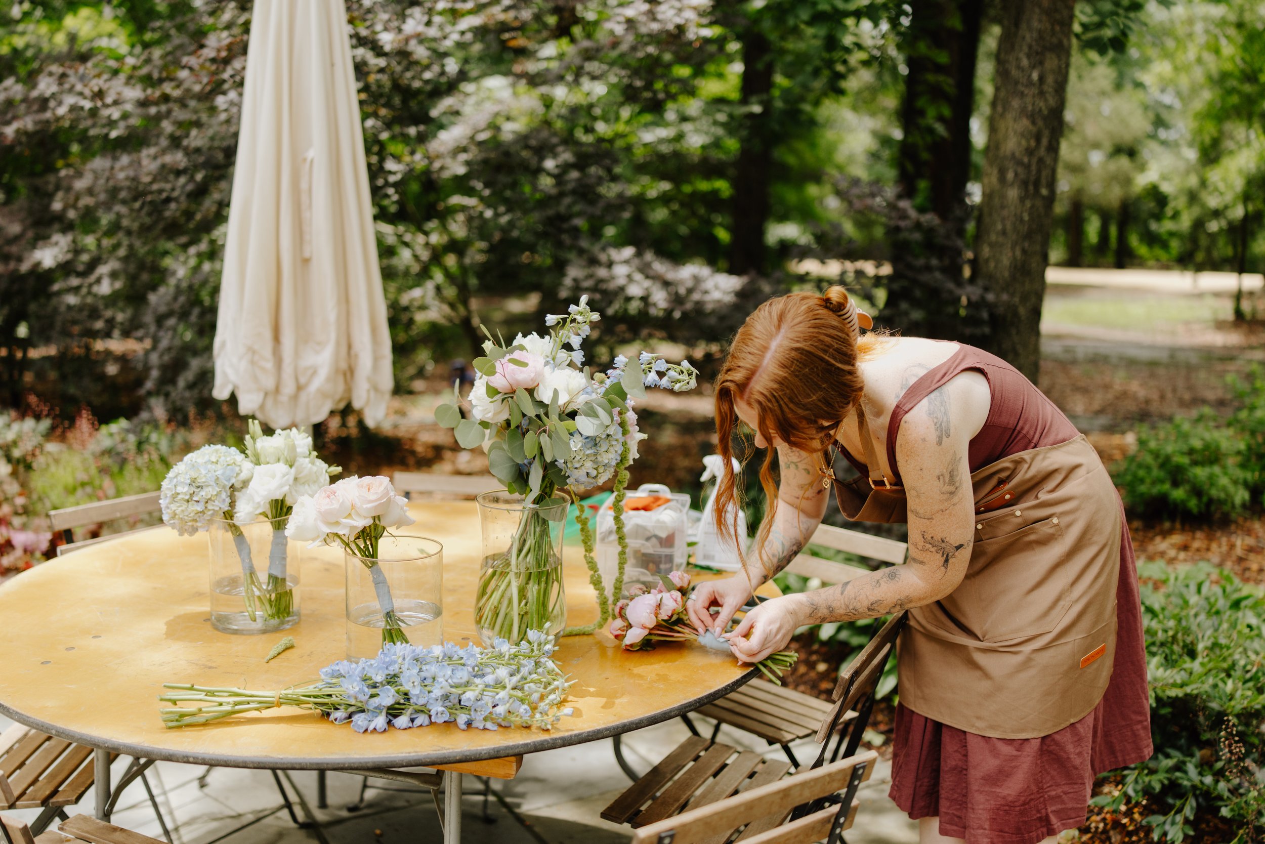 A person with red hair and tattoos, wearing a brown apron, arranging flowers on a table outdoors. The table has multiple vases filled with white and pink flowers, and some light blue flowers are on the table. There is a closed white umbrella and trees in the background.