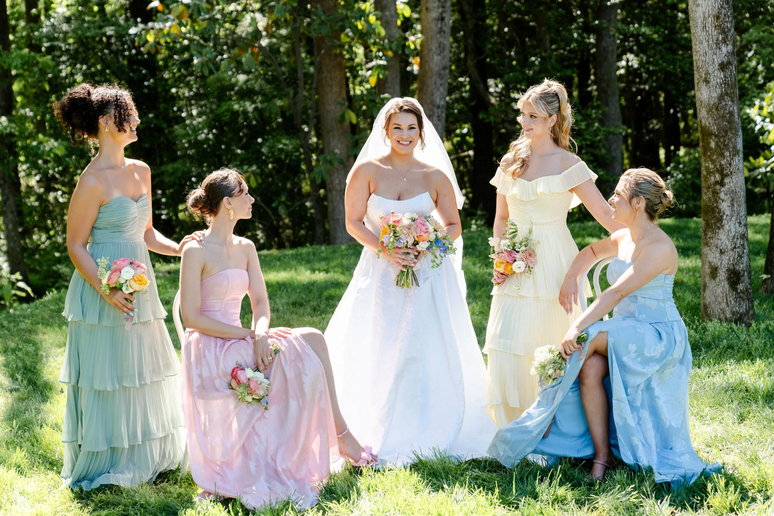 Group of women dressed in pastel colored dresses, with one woman in a white wedding dress holding a bouquet, outdoors in a wooded area.