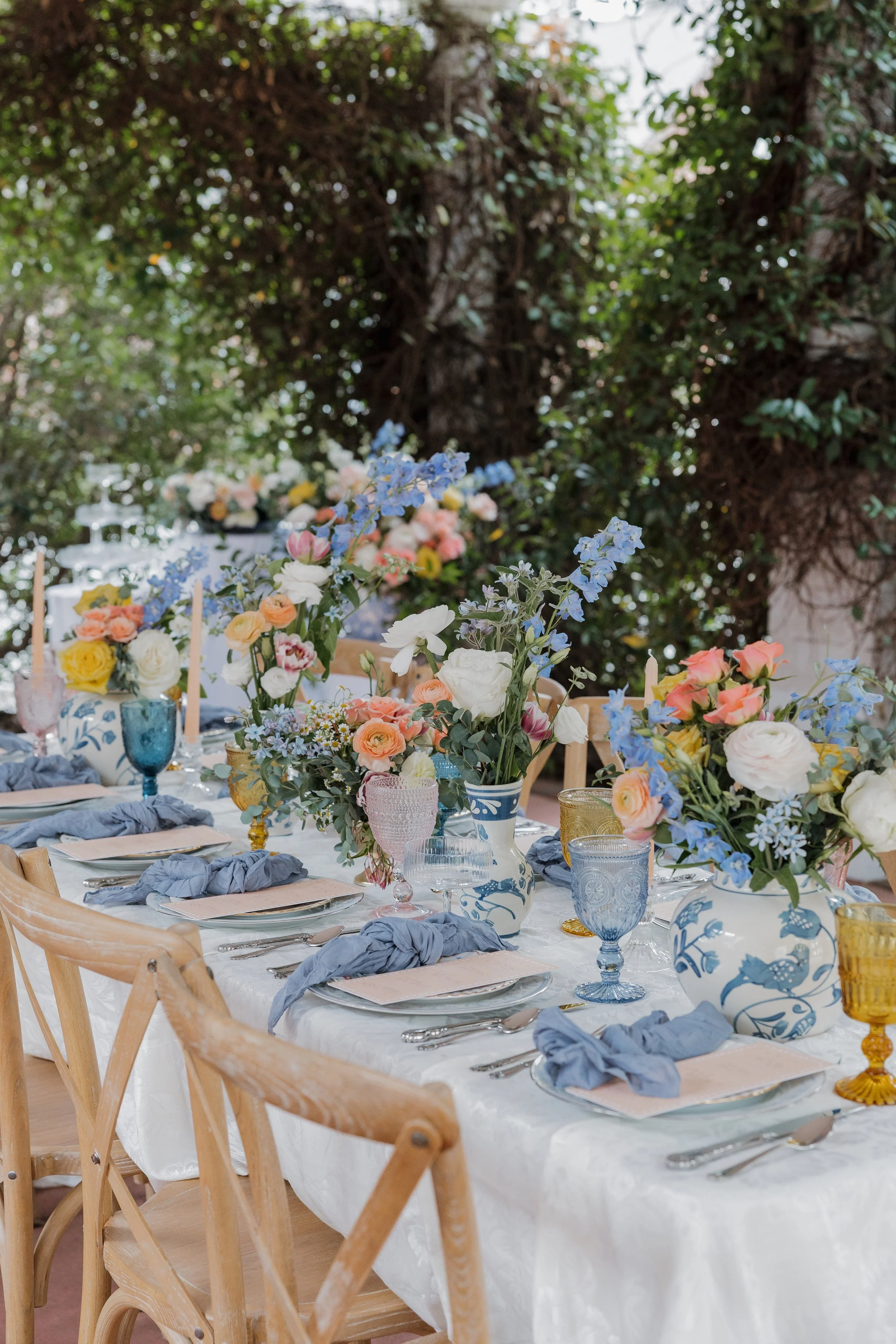A long dining table decorated with colorful flower arrangements in vases, set with plates, silverware, glasses, and napkins, outdoors under green trees for a celebration or gathering.