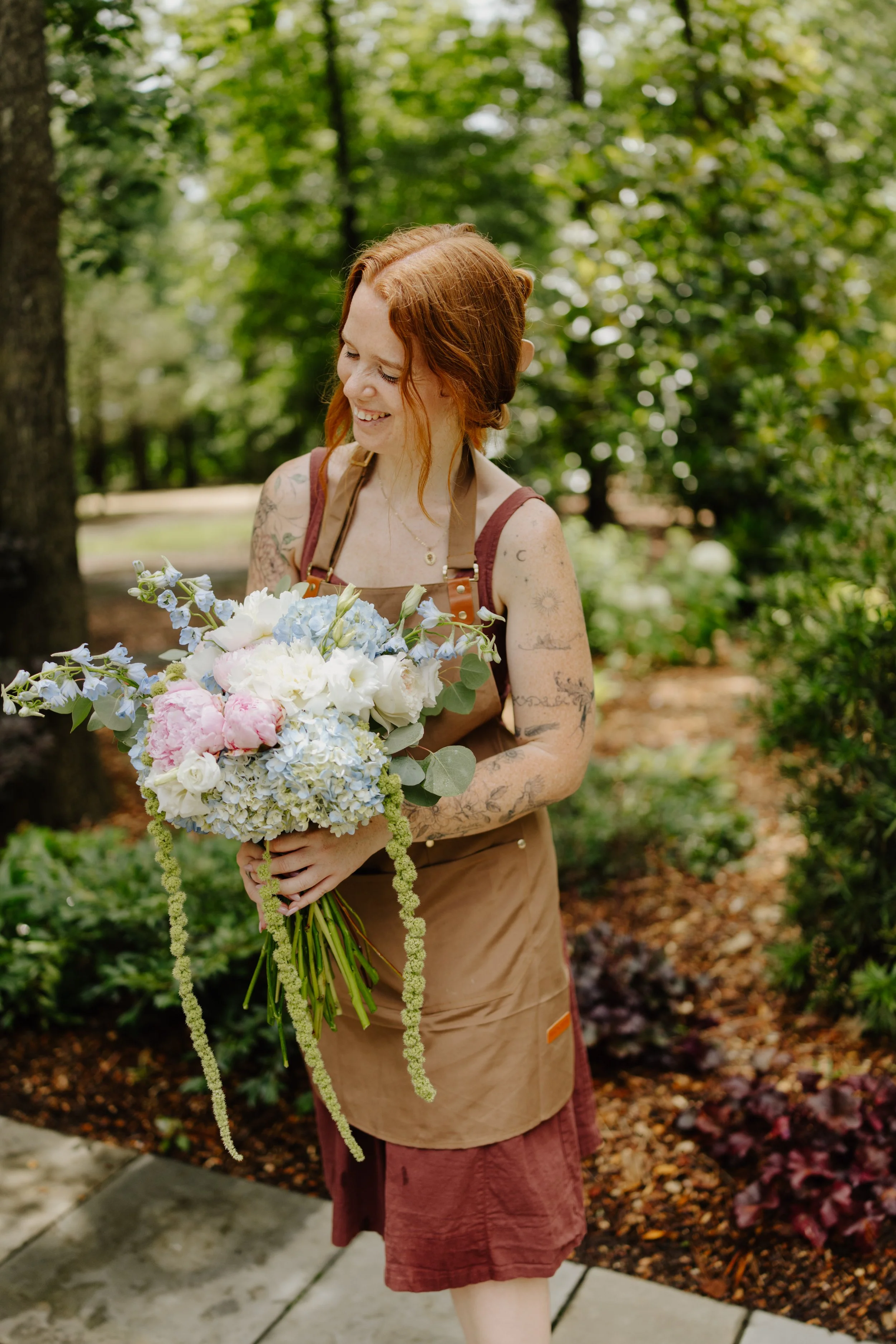 A young woman with tattoos and red hair, smiling and looking down, holding a bouquet of white, pink, and blue flowers, standing outdoors in a wooded area with green foliage in the background.
