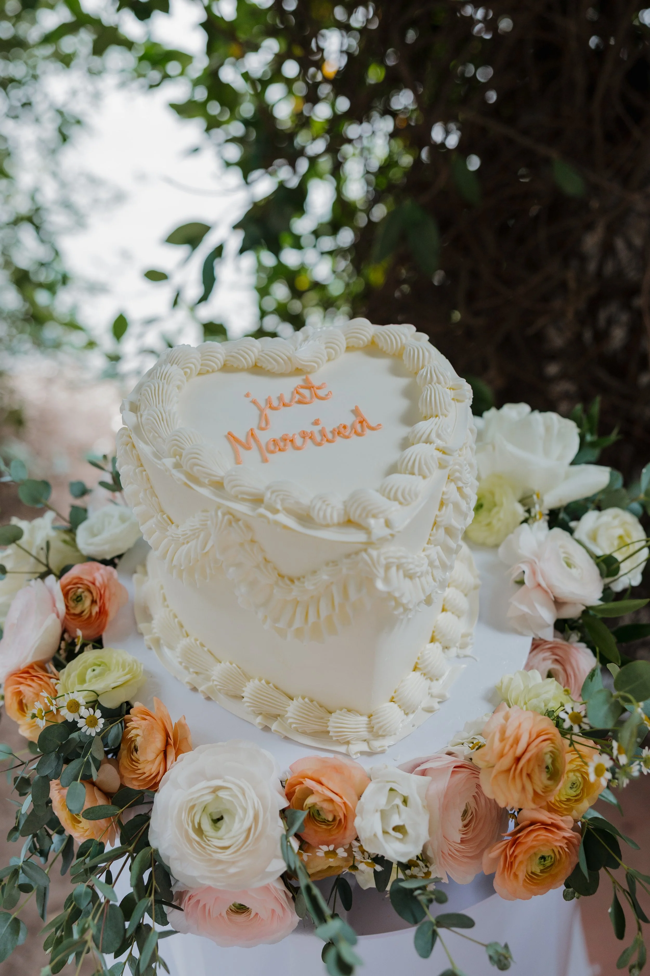 A wedding cake with white icing and decorative piping, surrounded by a colorful array of flowers including roses and daisies, with a blurred outdoor background.