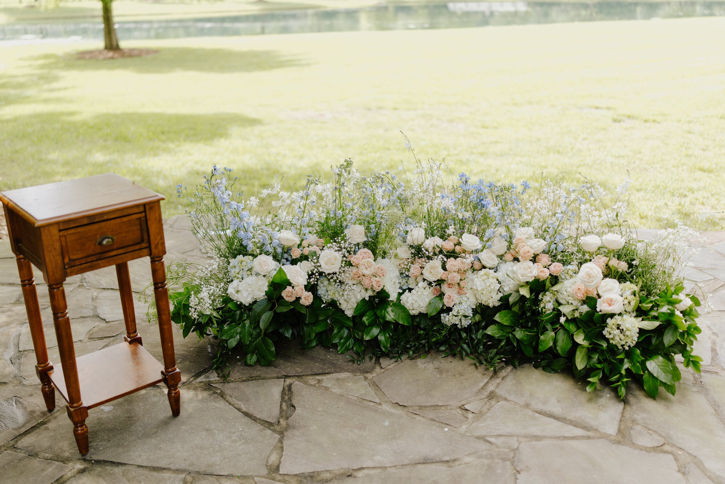 A wooden table next to a large floral arrangement of white and light pink roses, and light blue and white flowers, placed on a stone patio with a grassy area and a tree in the background.